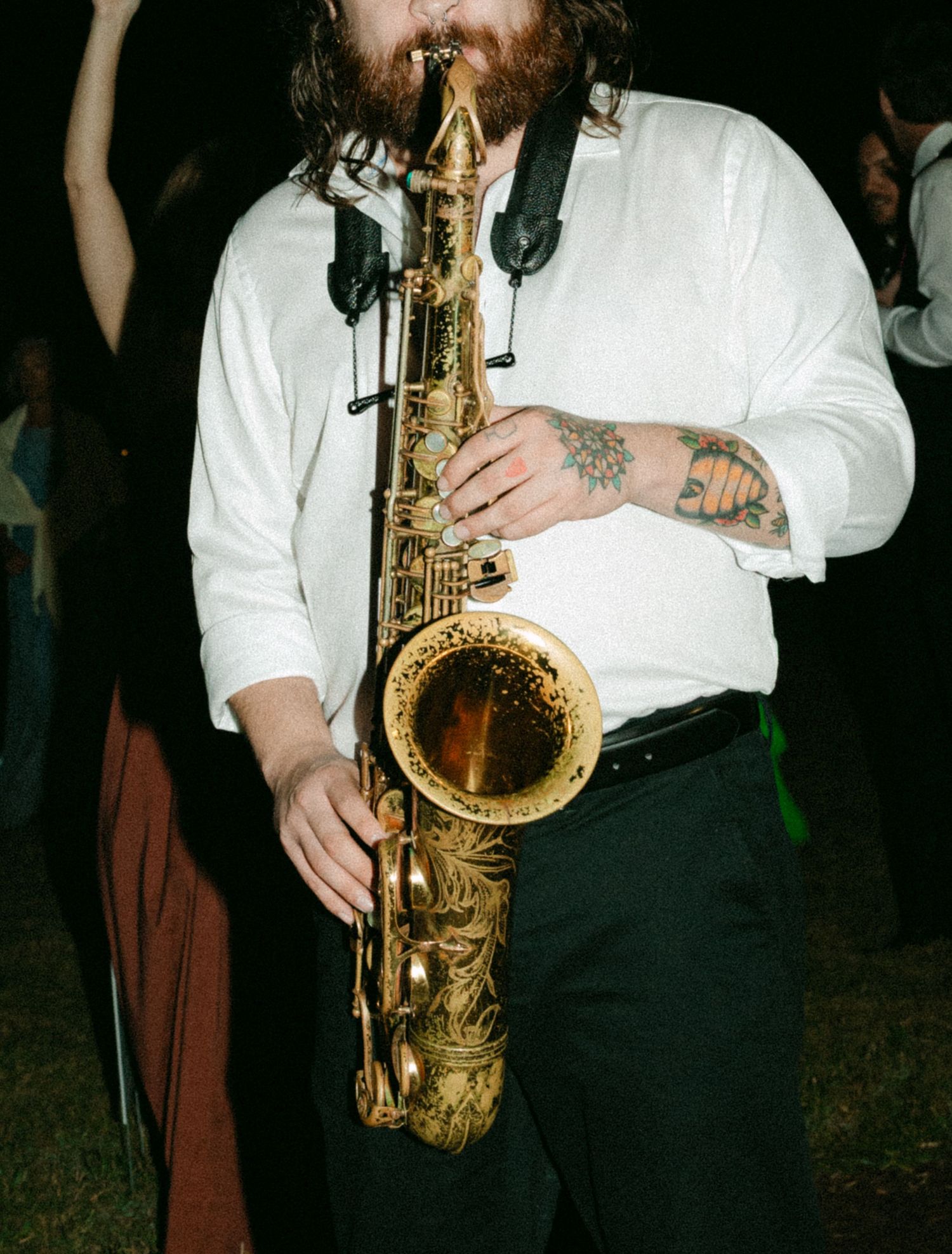 Bearded man with tattoos playing an ornate gold saxophone at an outdoor evening event.