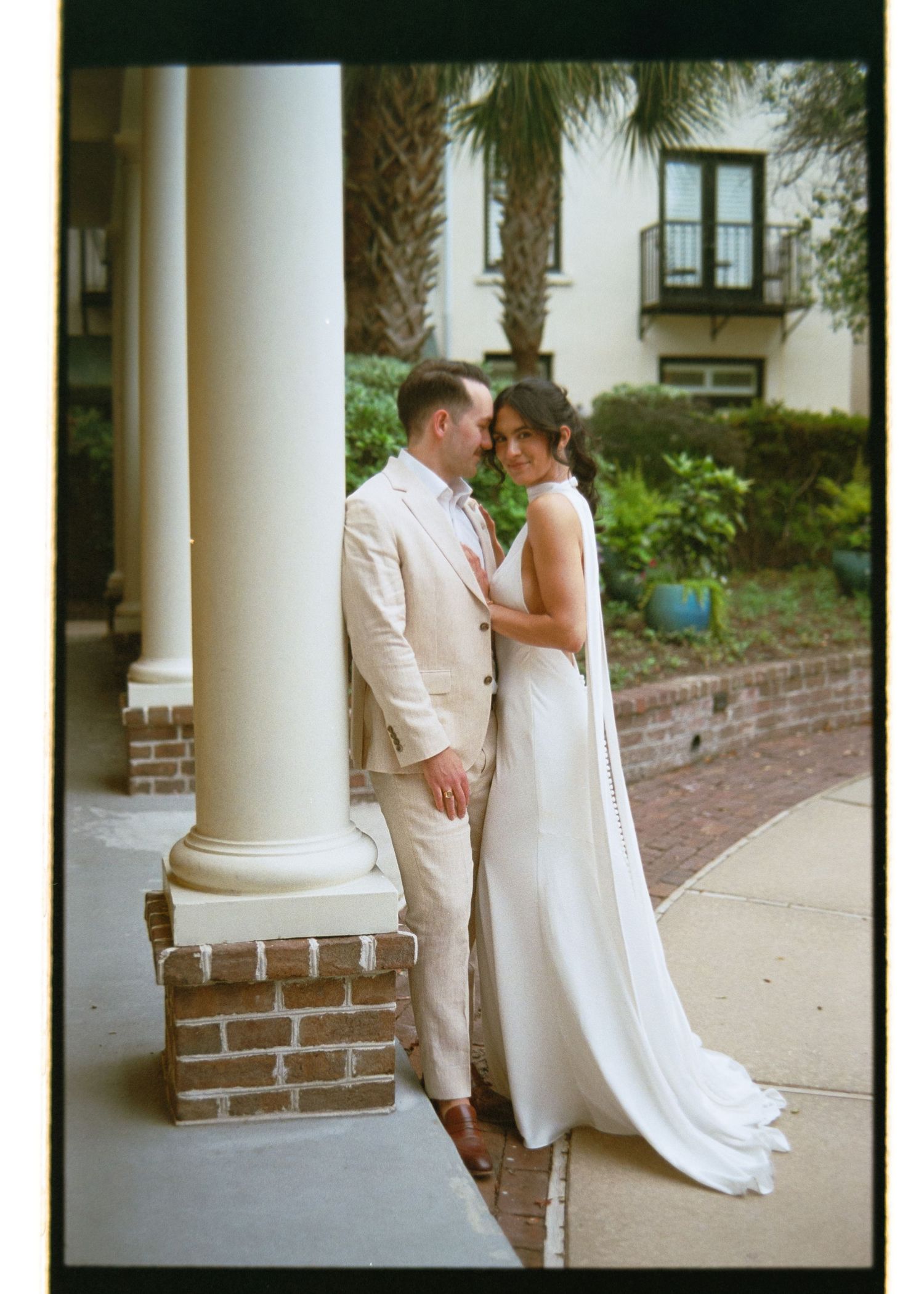 Bride and groom sharing an intimate moment beside white columns, bride wearing a sleek gown with long train.