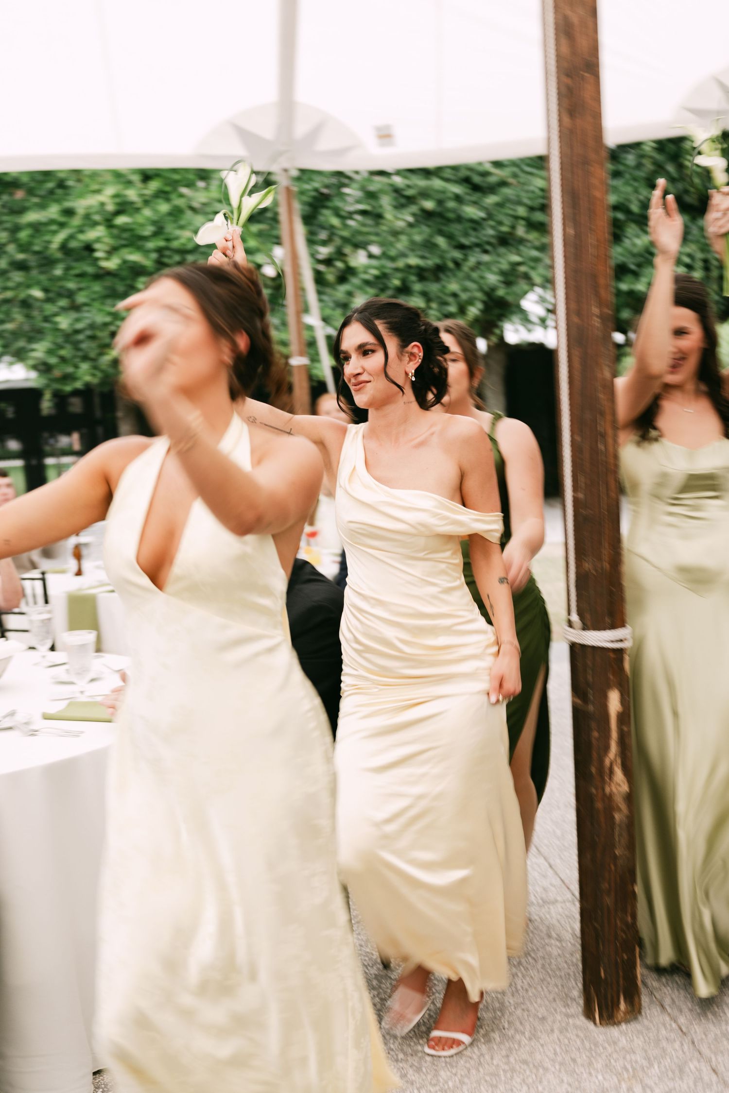 Bridesmaids in yellow satin gowns dancing joyfully at an outdoor wedding reception.
