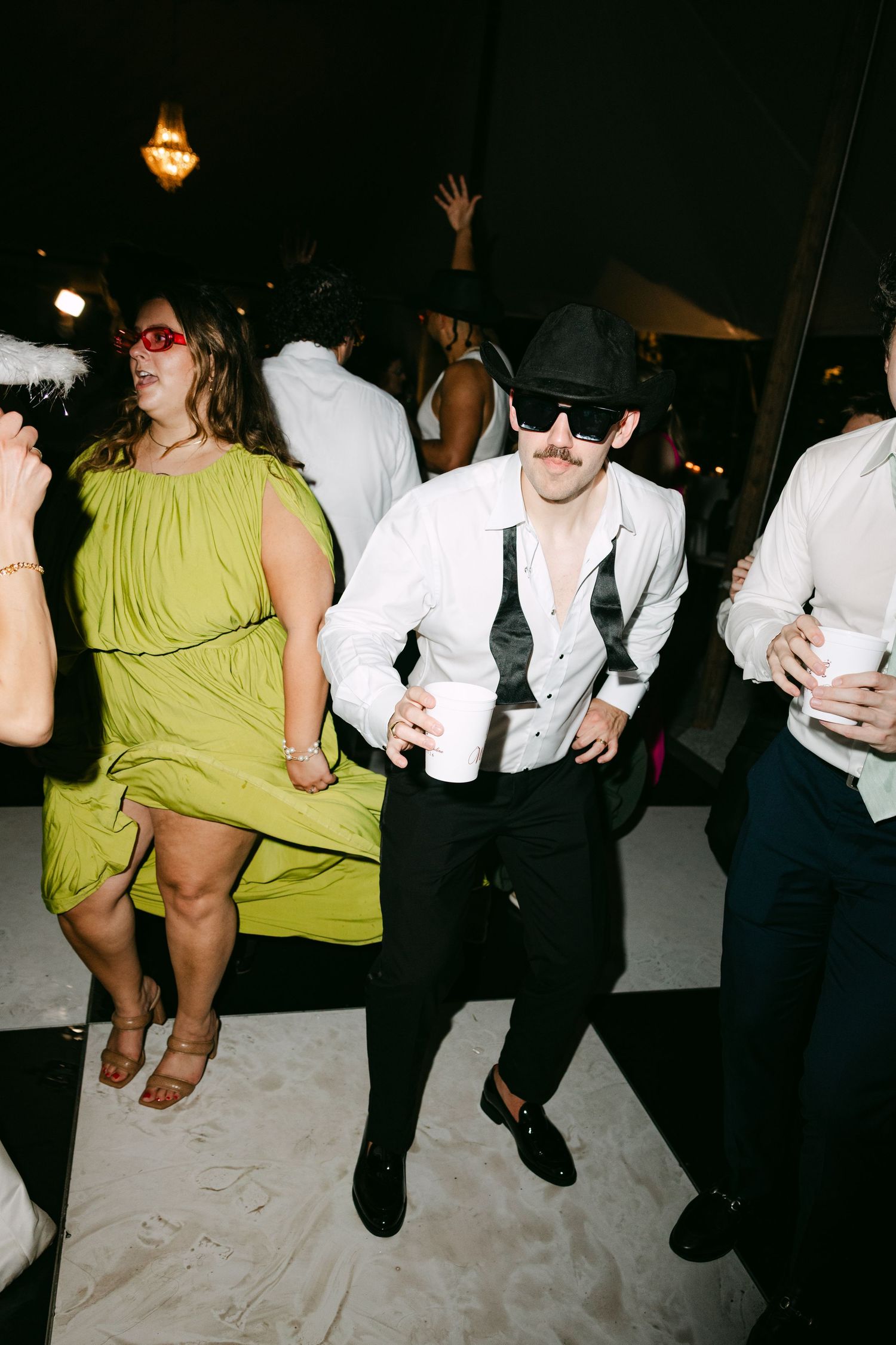 Wedding guest in green dress dancing with groom in black hat and sunglasses at reception.