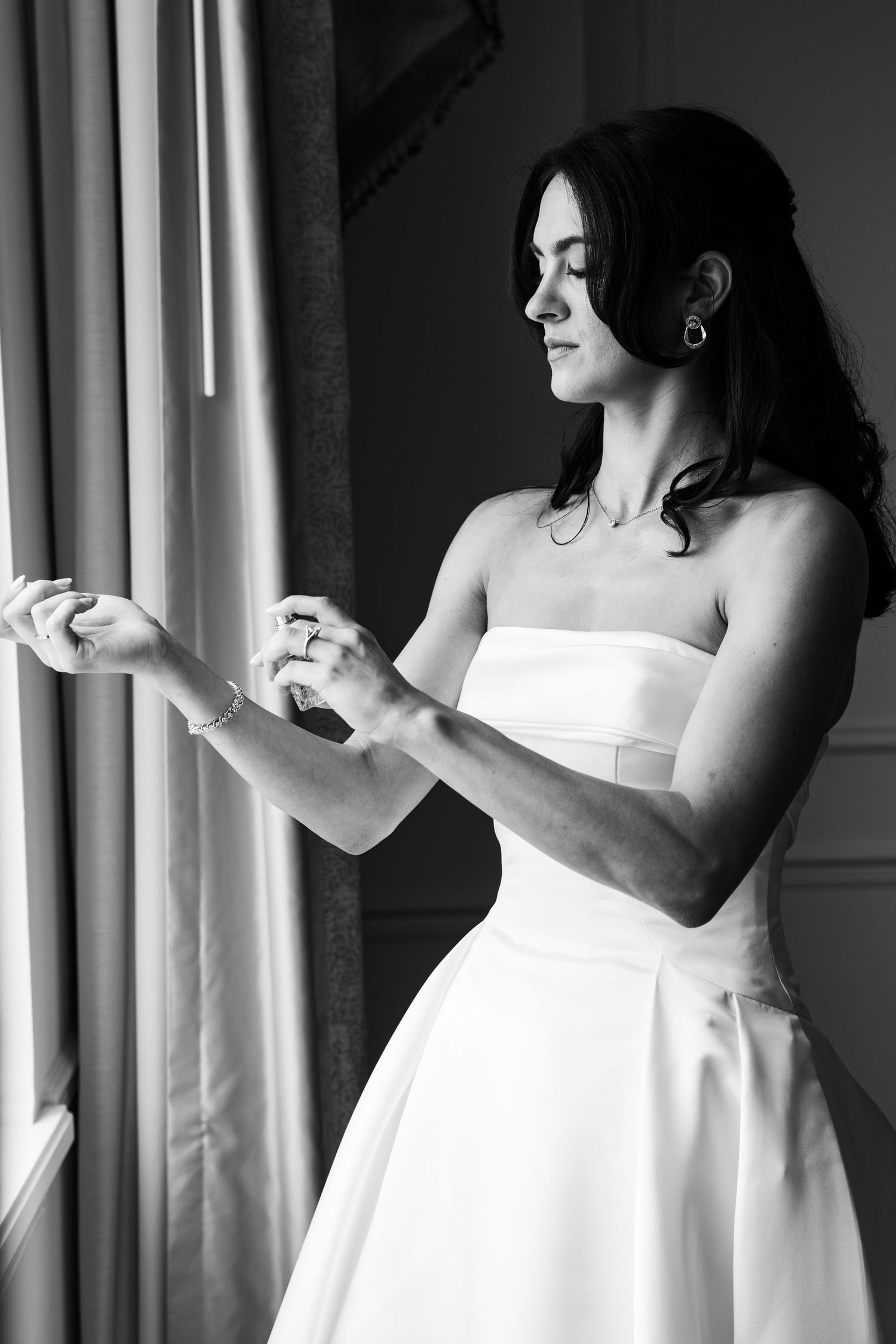 Black and white photo of a bride in a strapless gown adjusting her bracelet by a window.