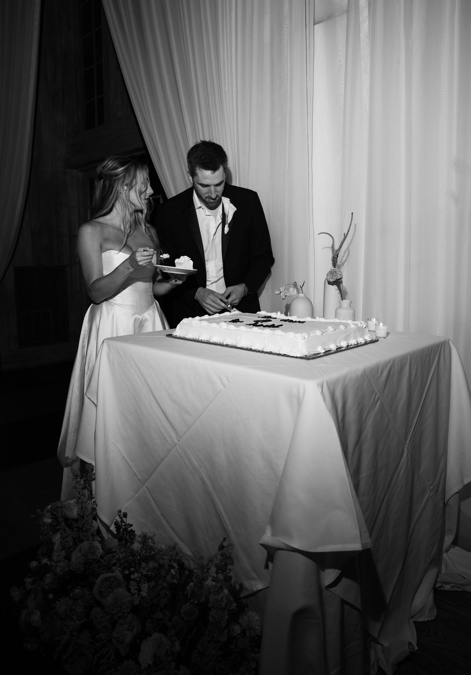 Black and white photo of bride and groom cutting wedding cake at elegant draped reception venue.
