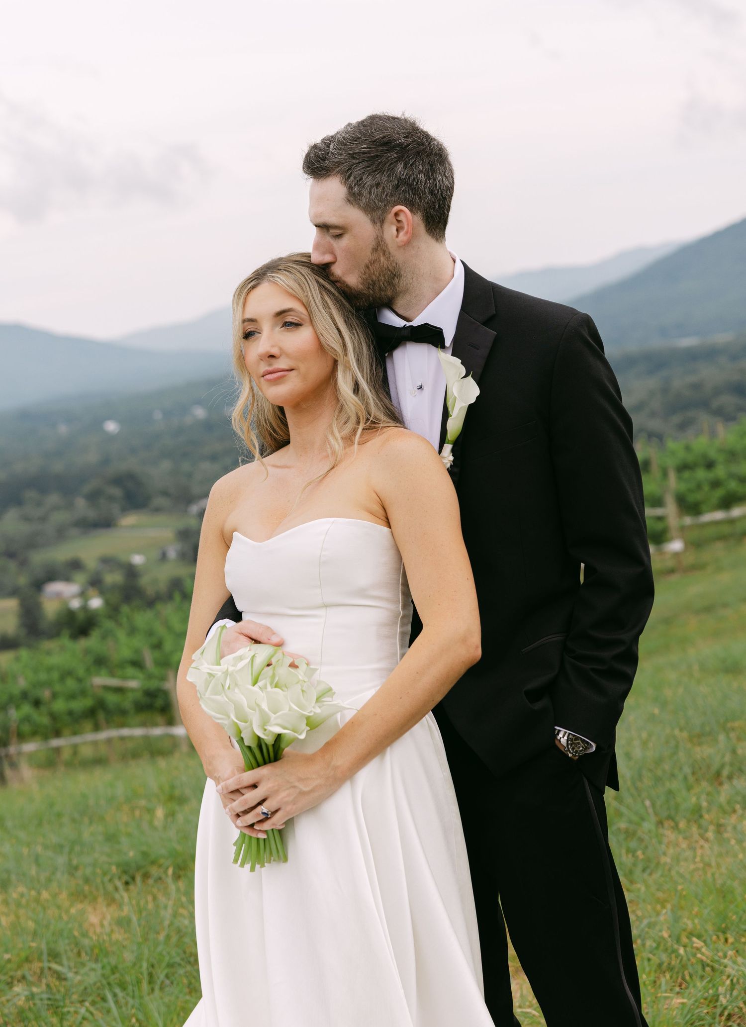 Bride and groom pose outdoors with mountain views, she holds white bouquet in elegant wedding attire.