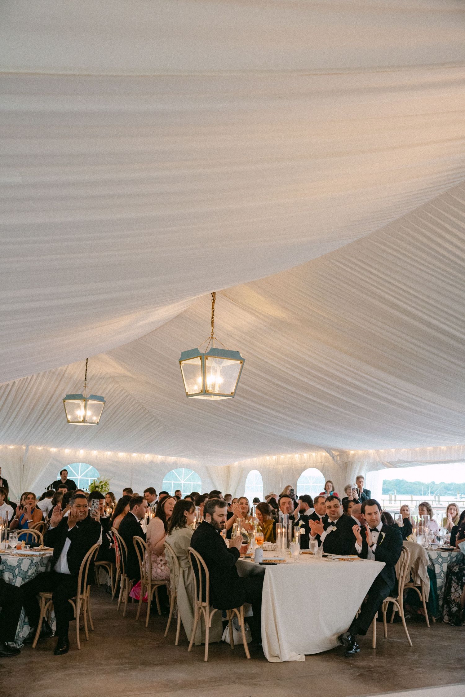 Elegant wedding reception under a white draped tent with lanterns, guests in formal attire seated at round tables.