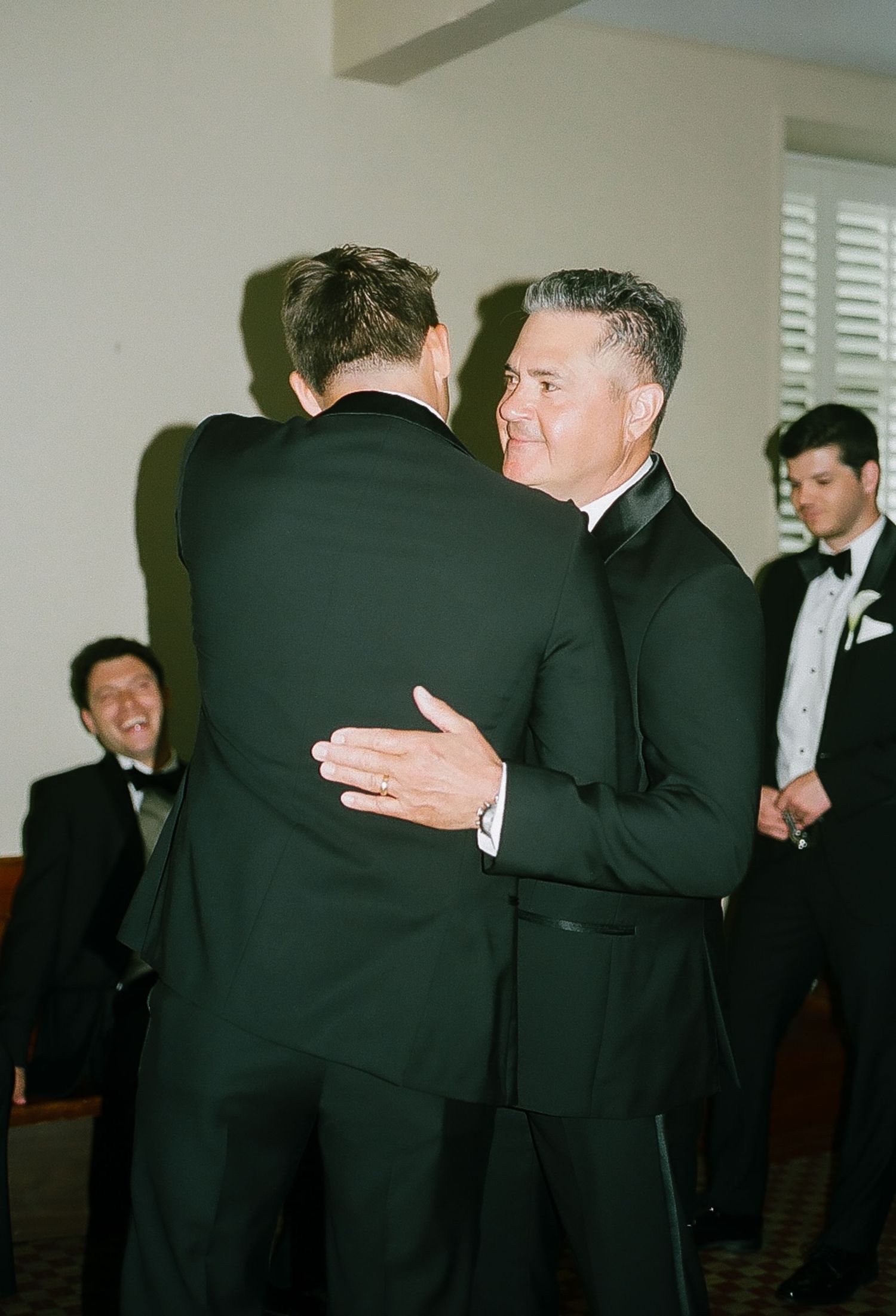 Groomsmen in black tuxedos share embraces during wedding preparations.