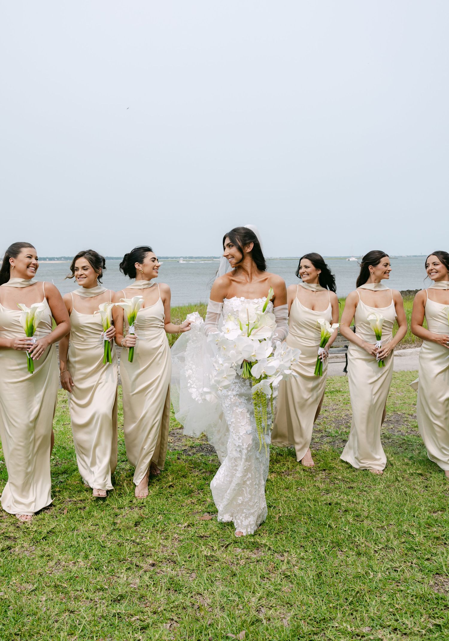 Bridesmaids in cream dresses holding bouquets form a playful line outdoors.