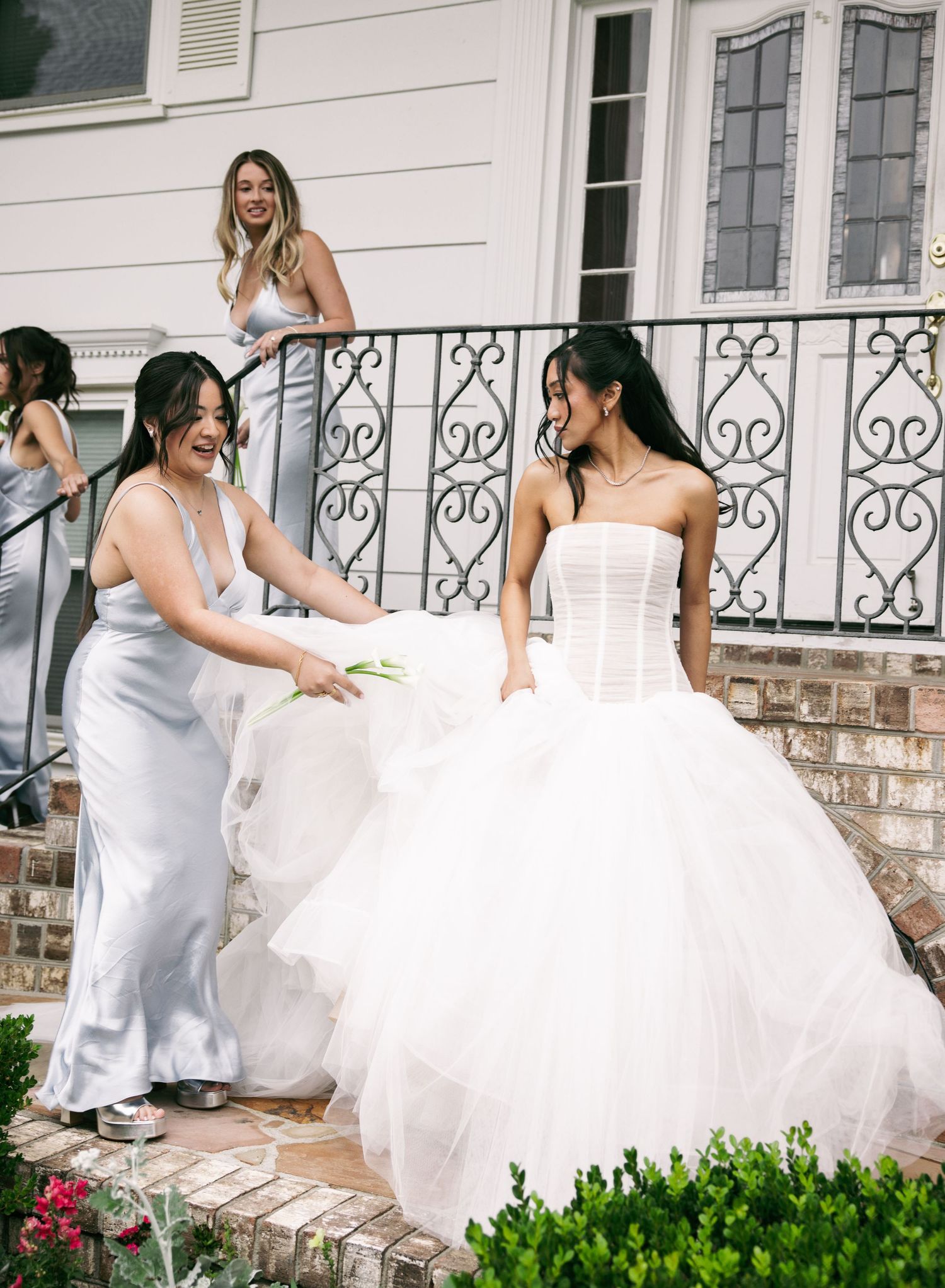 Bridesmaid helps bride in strapless corset ball gown on steps outside elegant home before wedding.