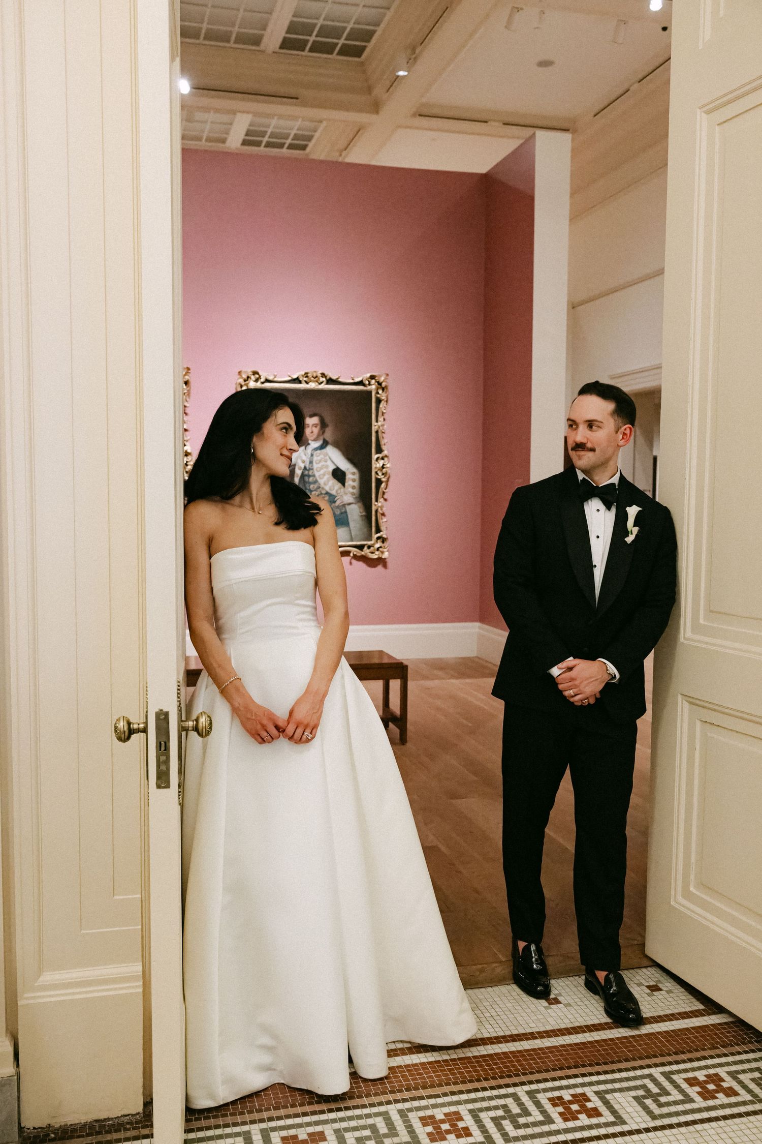Bride in strapless white gown and groom in tuxedo stand in museum doorway with pink gallery wall behind.