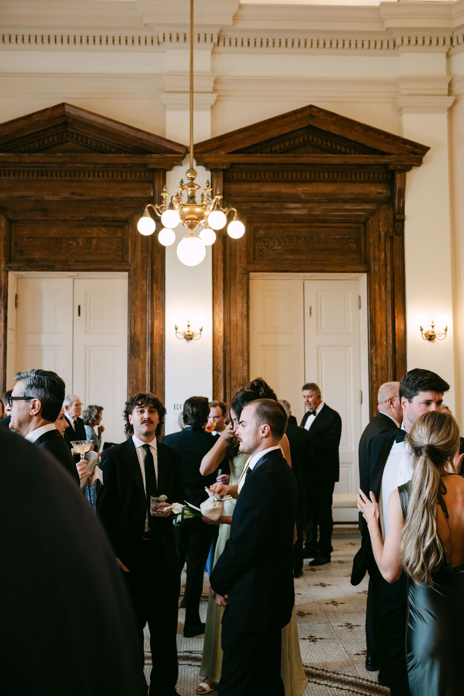 Elegant wedding cocktail hour in a grand ballroom with guests in formal attire mingling under ornate chandeliers.