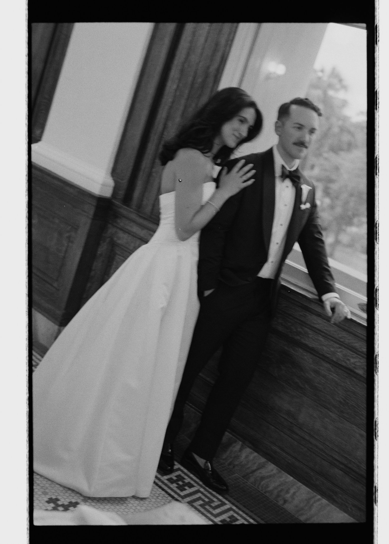 Black and white film photo of bride and groom posing on elegant venue balcony.