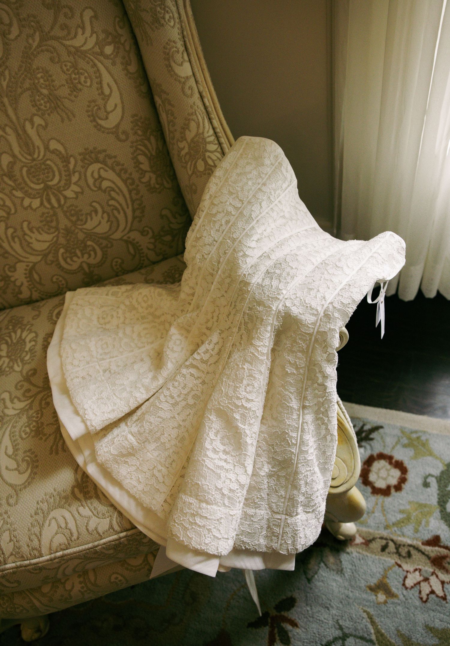 Elegant strapless white lace wedding dress draped over ornate vintage armchair near window.