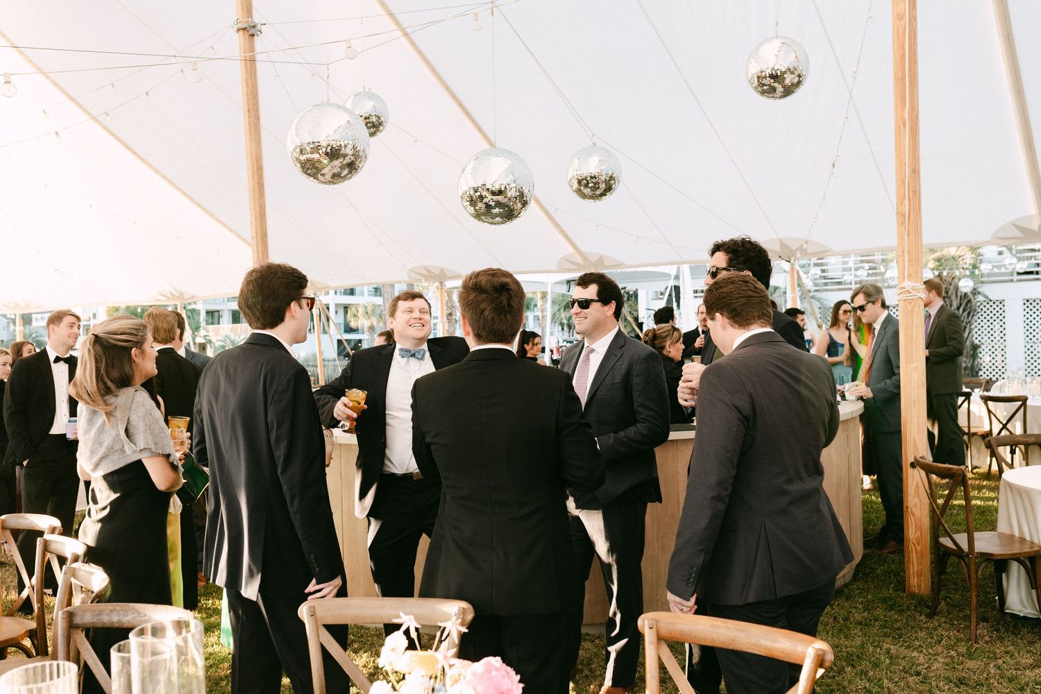 Wedding guests mingle under white sailcloth tent with hanging disco balls during outdoor reception cocktail hour.