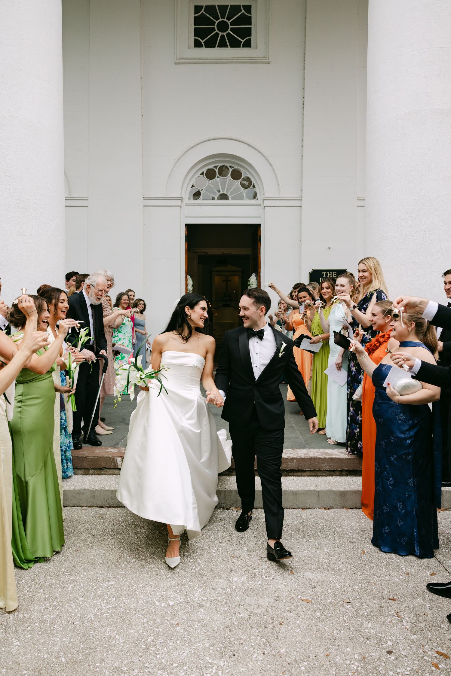 Newlywed couple exits church holding hands, greeted by cheering guests in colorful attire on sunny wedding day.