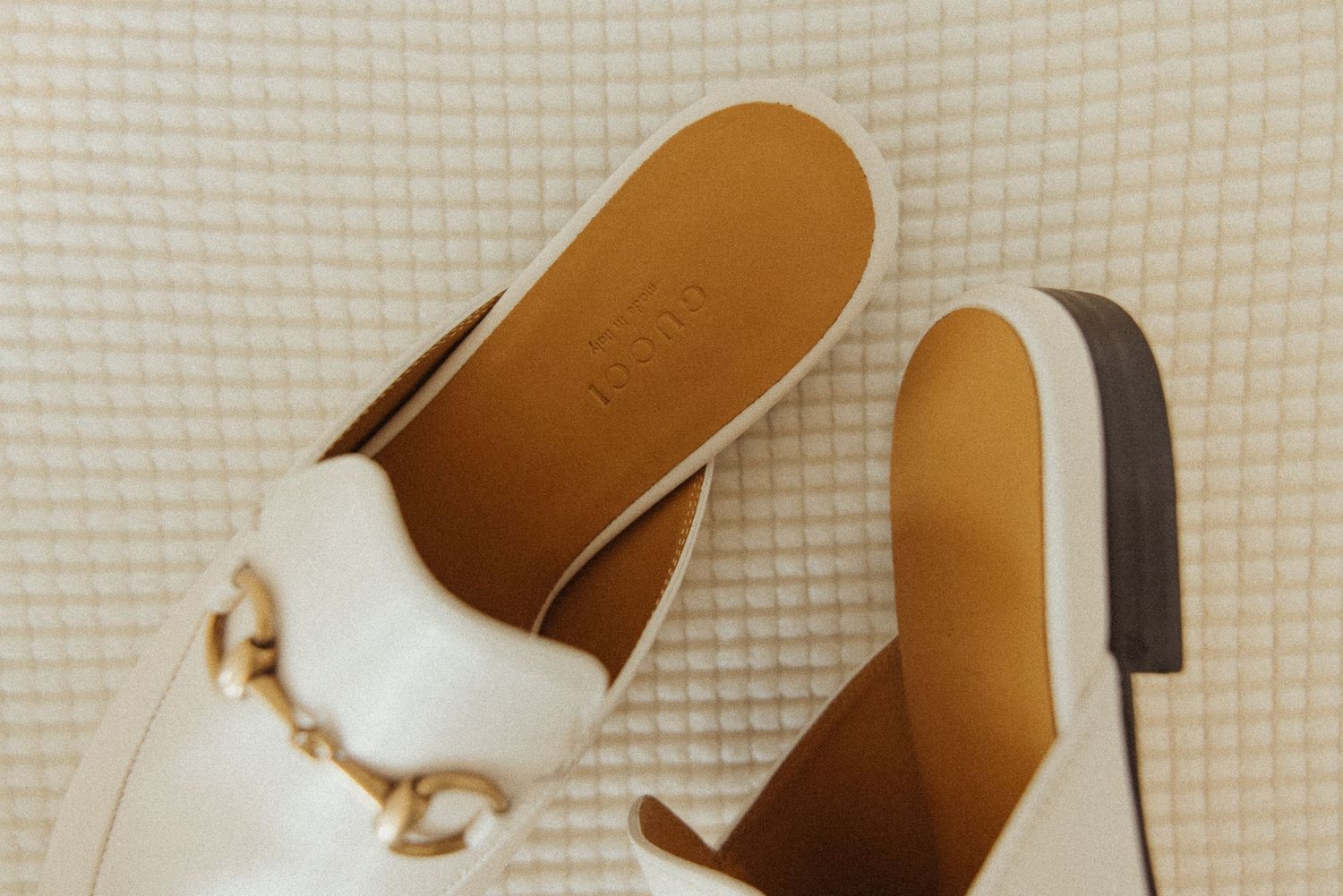 White leather mule slides with gold chain hardware detail and tan insole, displayed on textured background.
