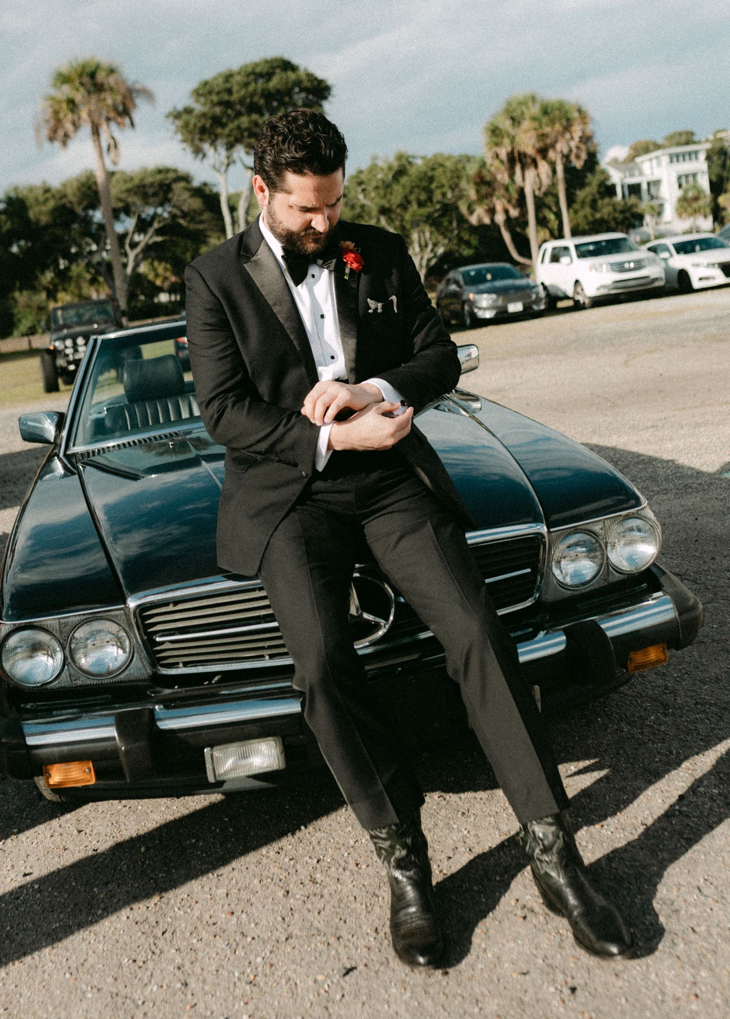 Dapper groom in black tuxedo leaning against vintage black convertible car outdoors.
