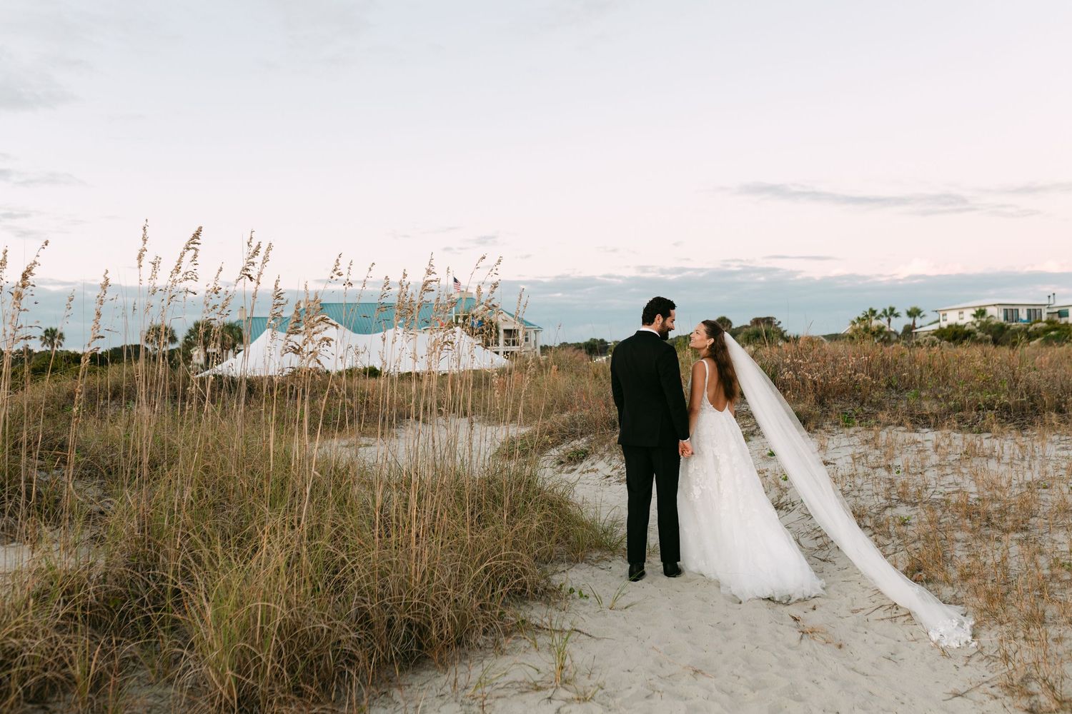Bride and groom stand together on sandy beach path with sea oats, facing a coastal wedding venue at dusk.