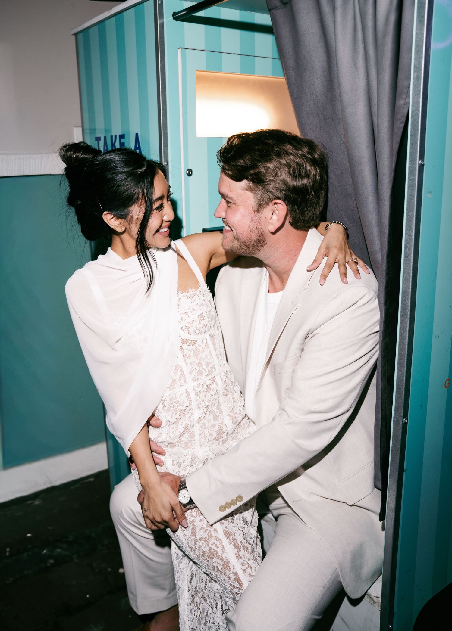 Couple in white outfits smiling and embracing inside a photo booth with teal striped walls.