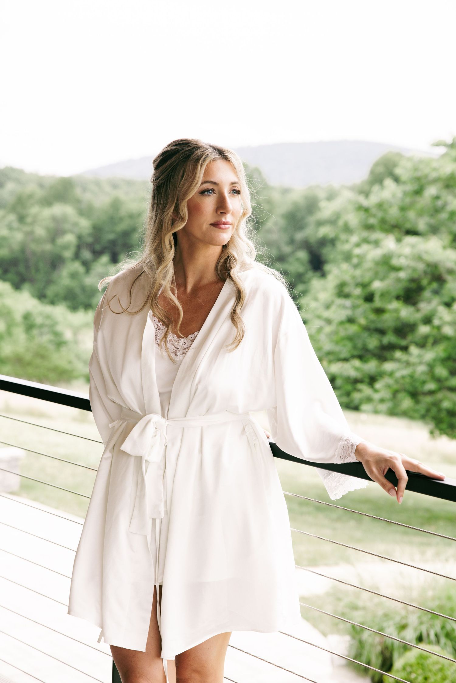 Blonde bride in white silk robe stands on balcony with lush green mountain views behind her.
