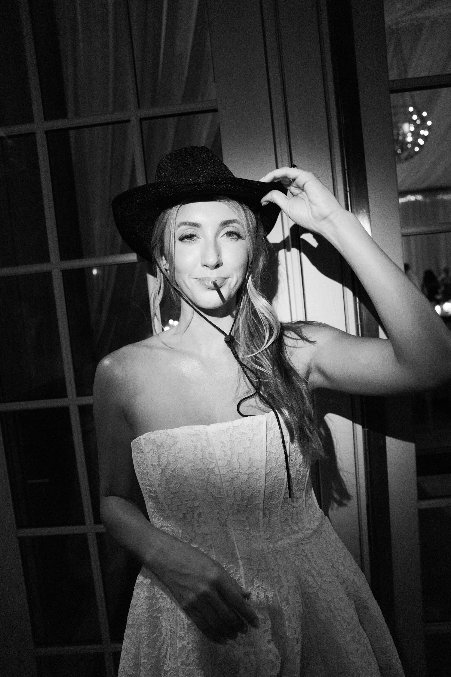 Black and white photo of a woman in a strapless dress and cowboy hat posing indoors at a party.