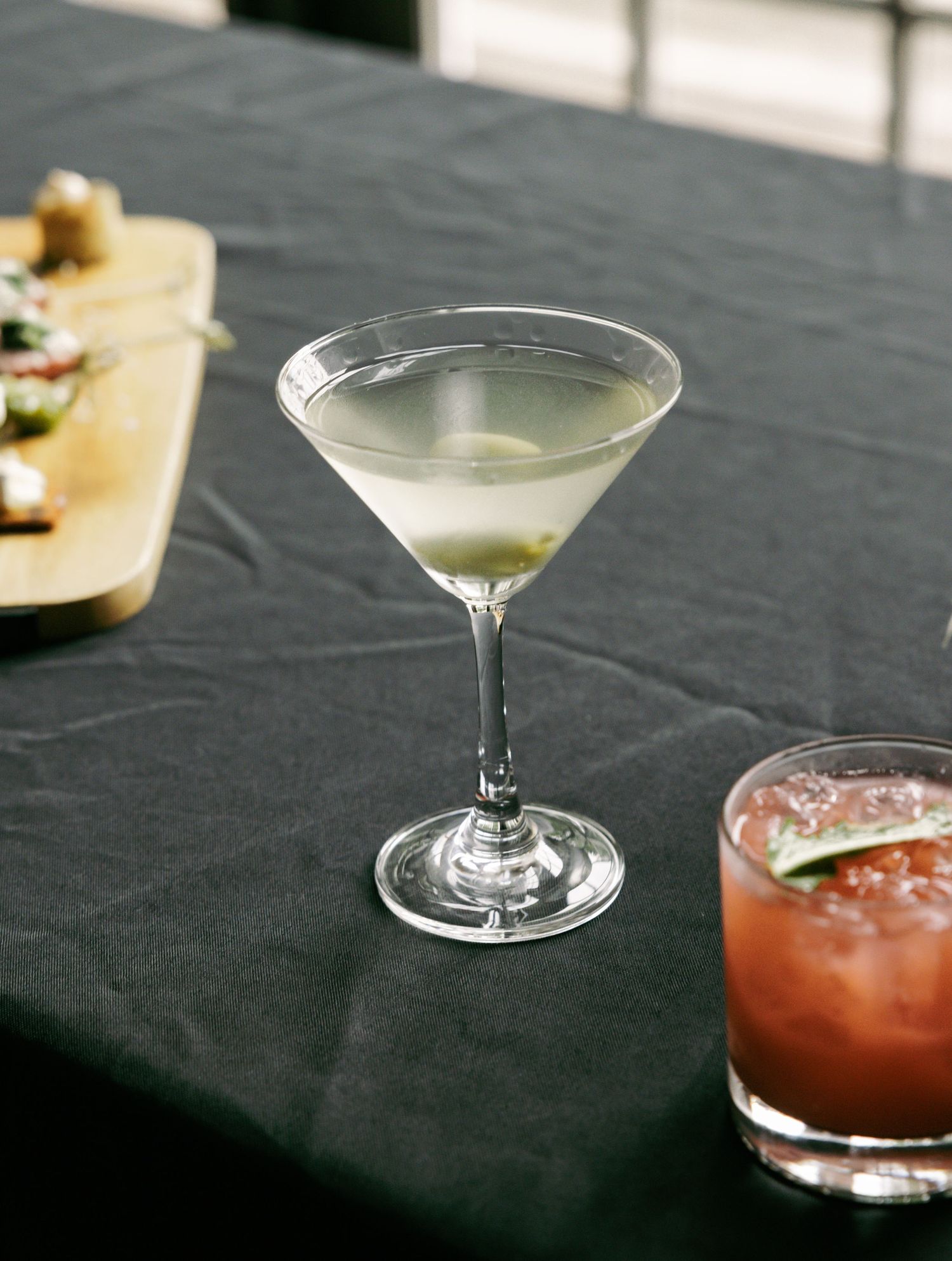A classic dirty martini with olive garnish beside a red cocktail on a black linen tablecloth at an elegant event.