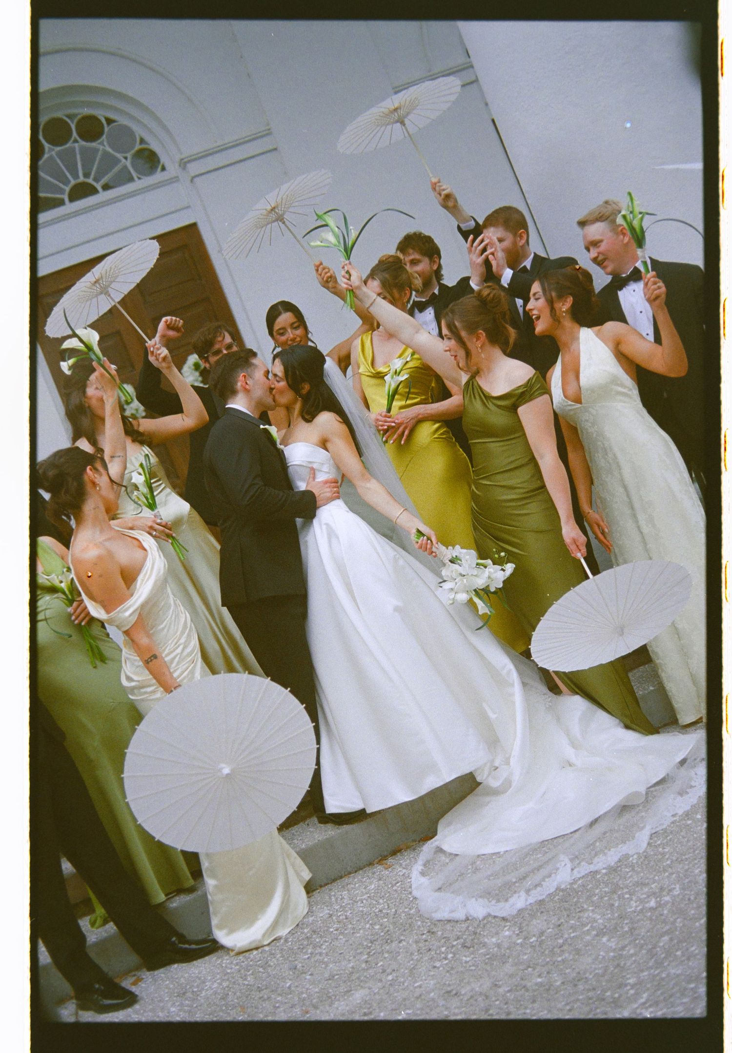 Colorful film photo of wedding party cheering with parasols as newlyweds kiss outside white church.