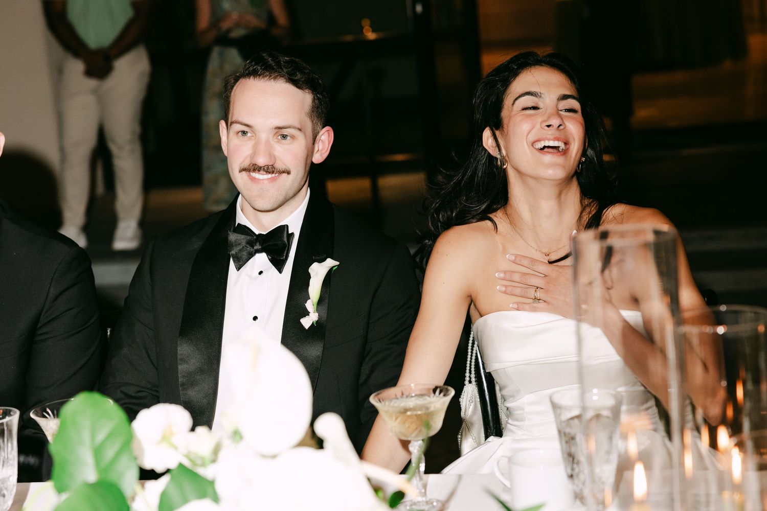 Bride and groom laugh together at sweetheart table during elegant candlelit wedding reception.