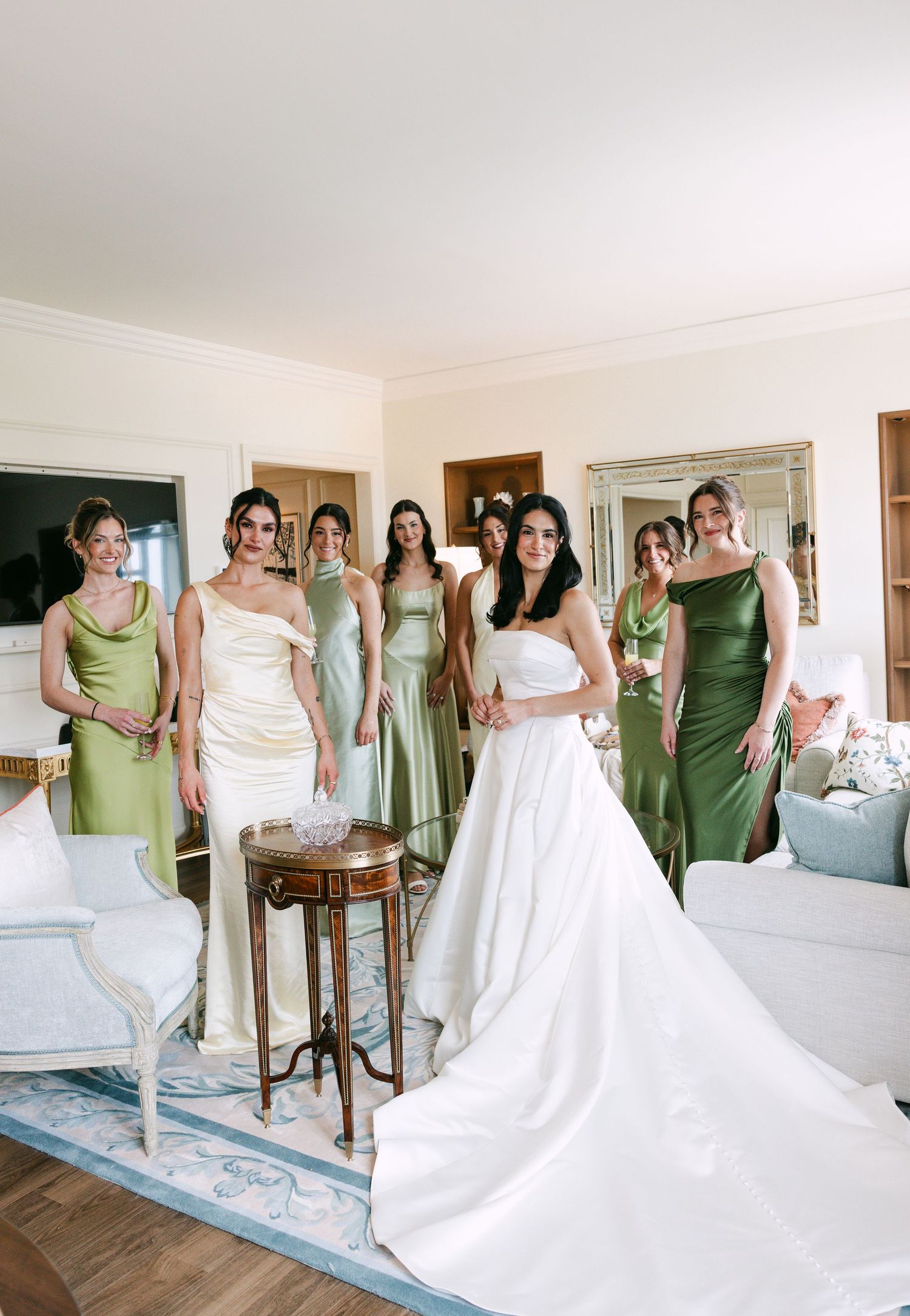 Bride in white gown poses with bridesmaids in sage green satin dresses in an elegant room before the wedding ceremony.