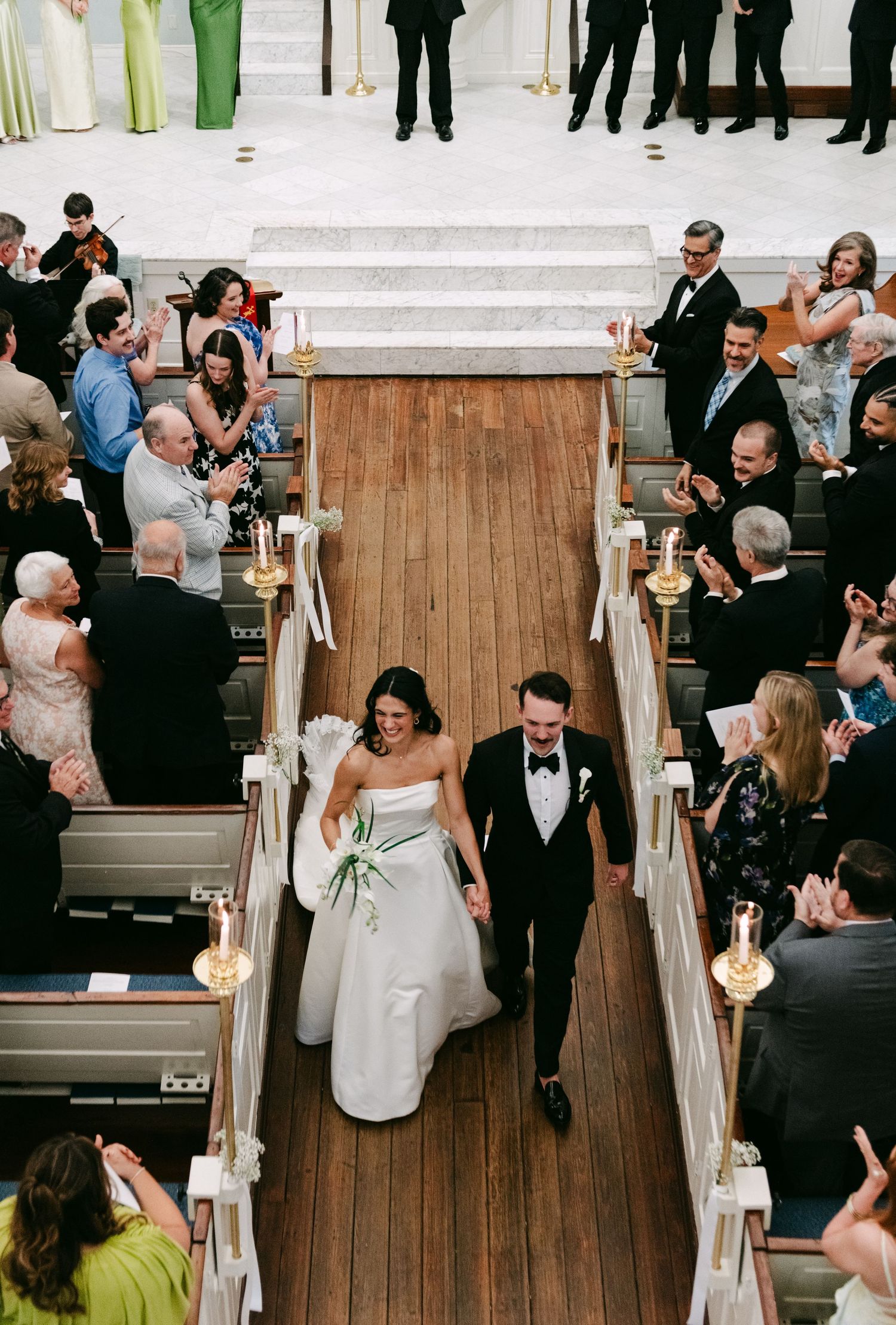 Joyful bride and groom walk down church aisle as guests applaud during wedding ceremony recessional.