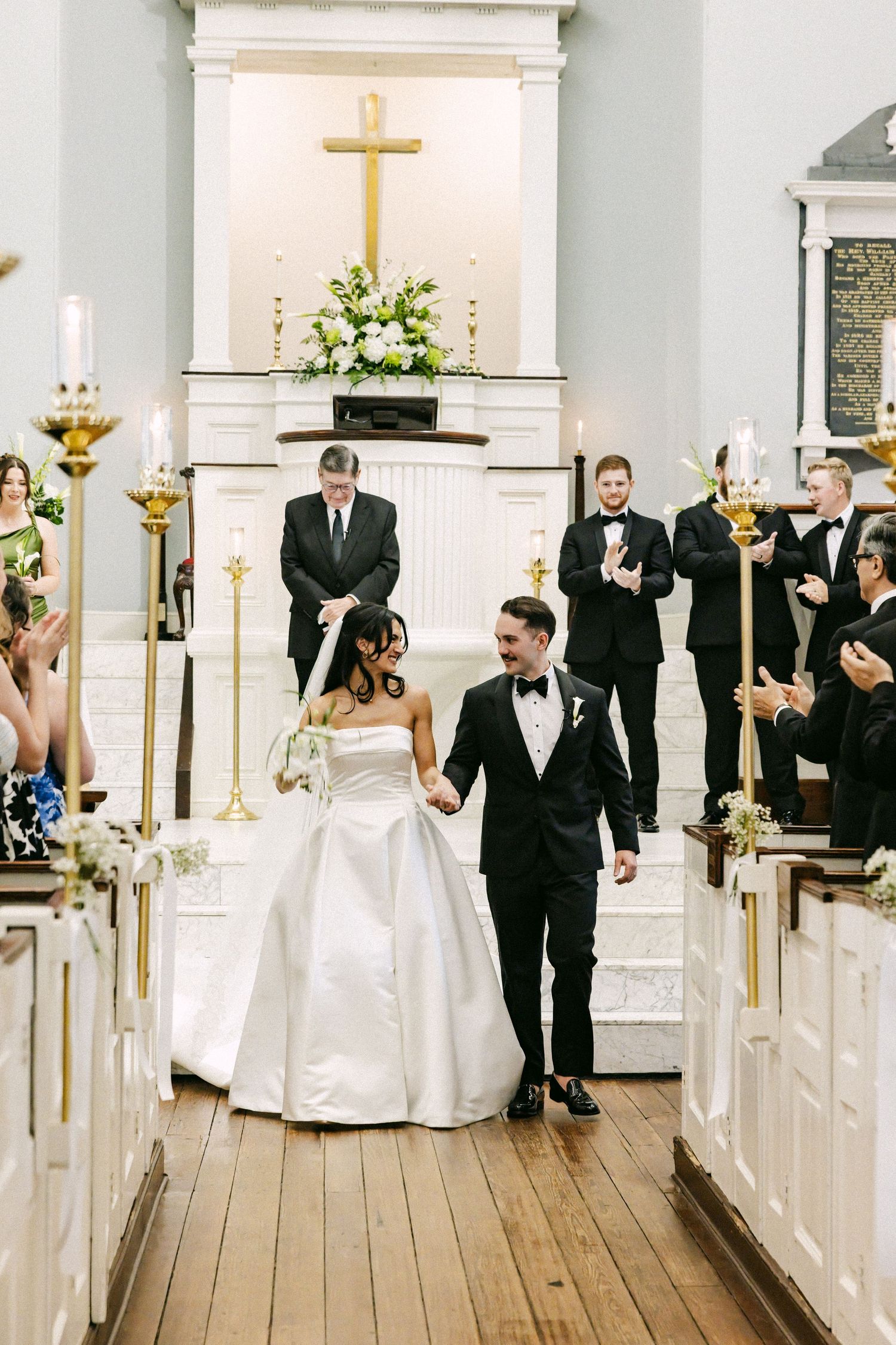 Bride and groom walk down church aisle after wedding ceremony, surrounded by guests applauding in elegant white chapel.