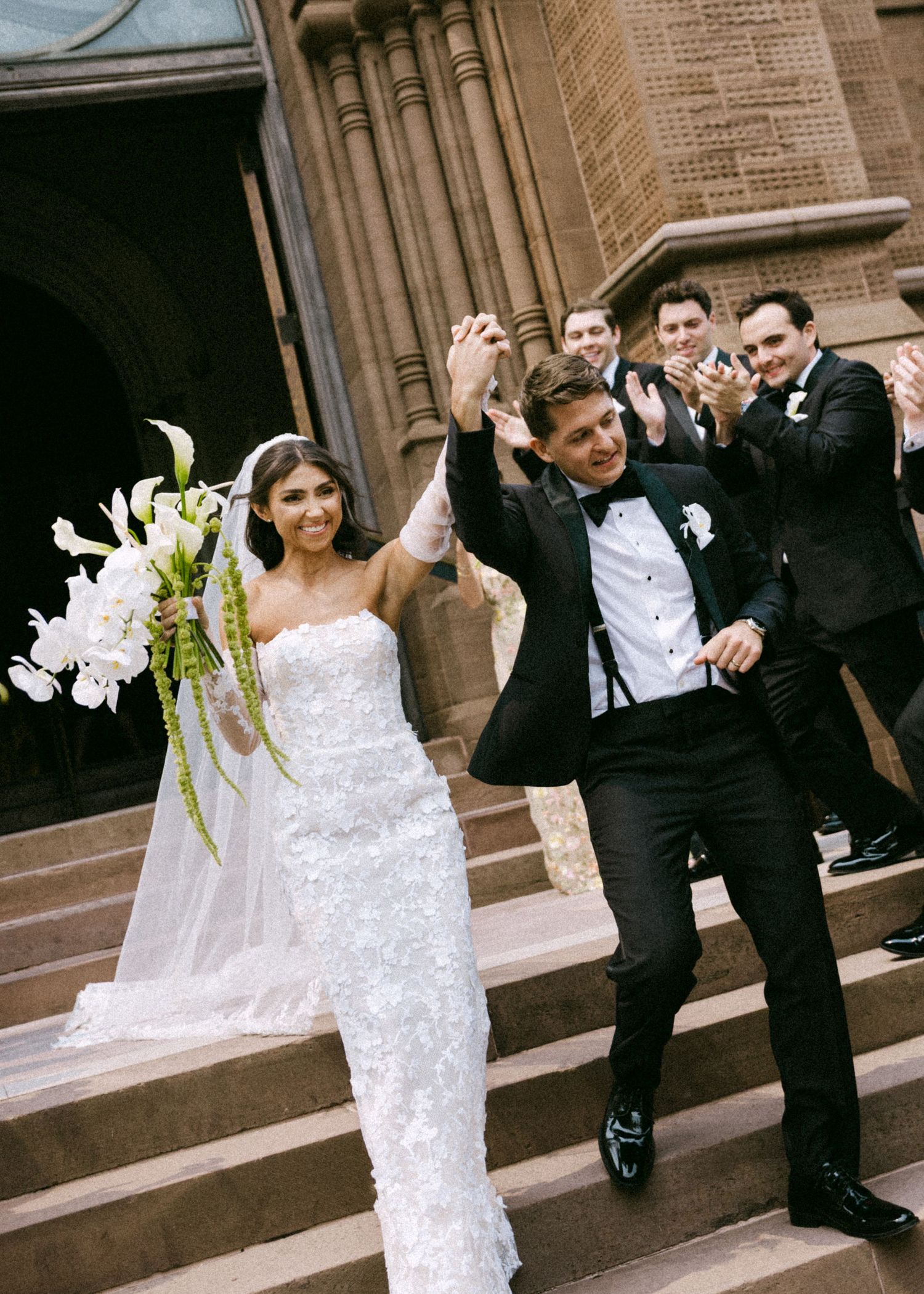 A bride in a white lace gown and groom in black tuxedo pose playfully outside a church.