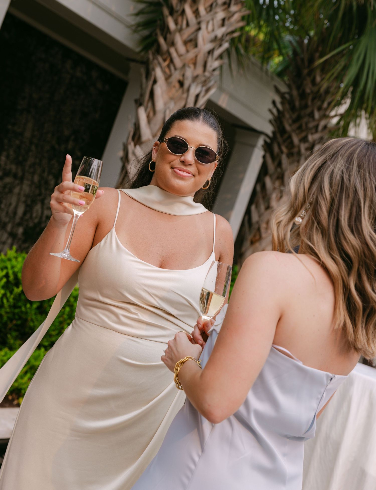 Wedding guests in light colored dresses toast with champagne glasses beside tropical palm trees.