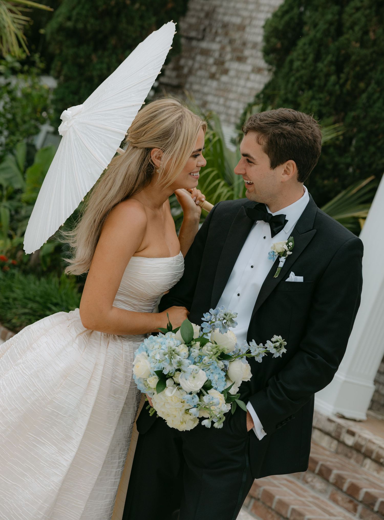 Wedding couple poses with white parasol in garden setting.