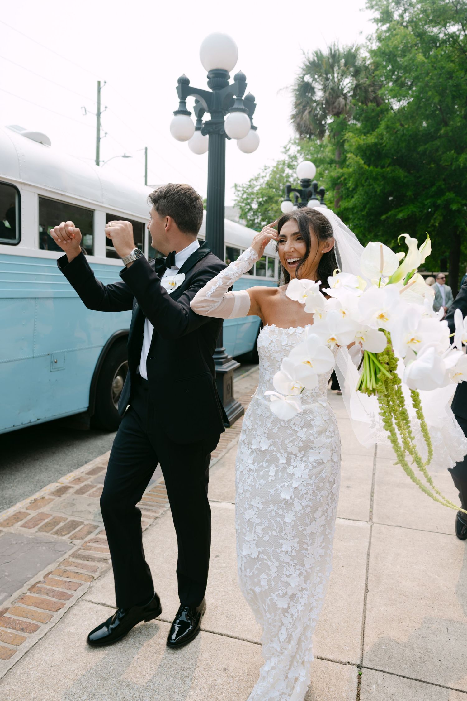 Wedding couple and attendants dance joyfully on a sidewalk with white flowers and formal attire.
