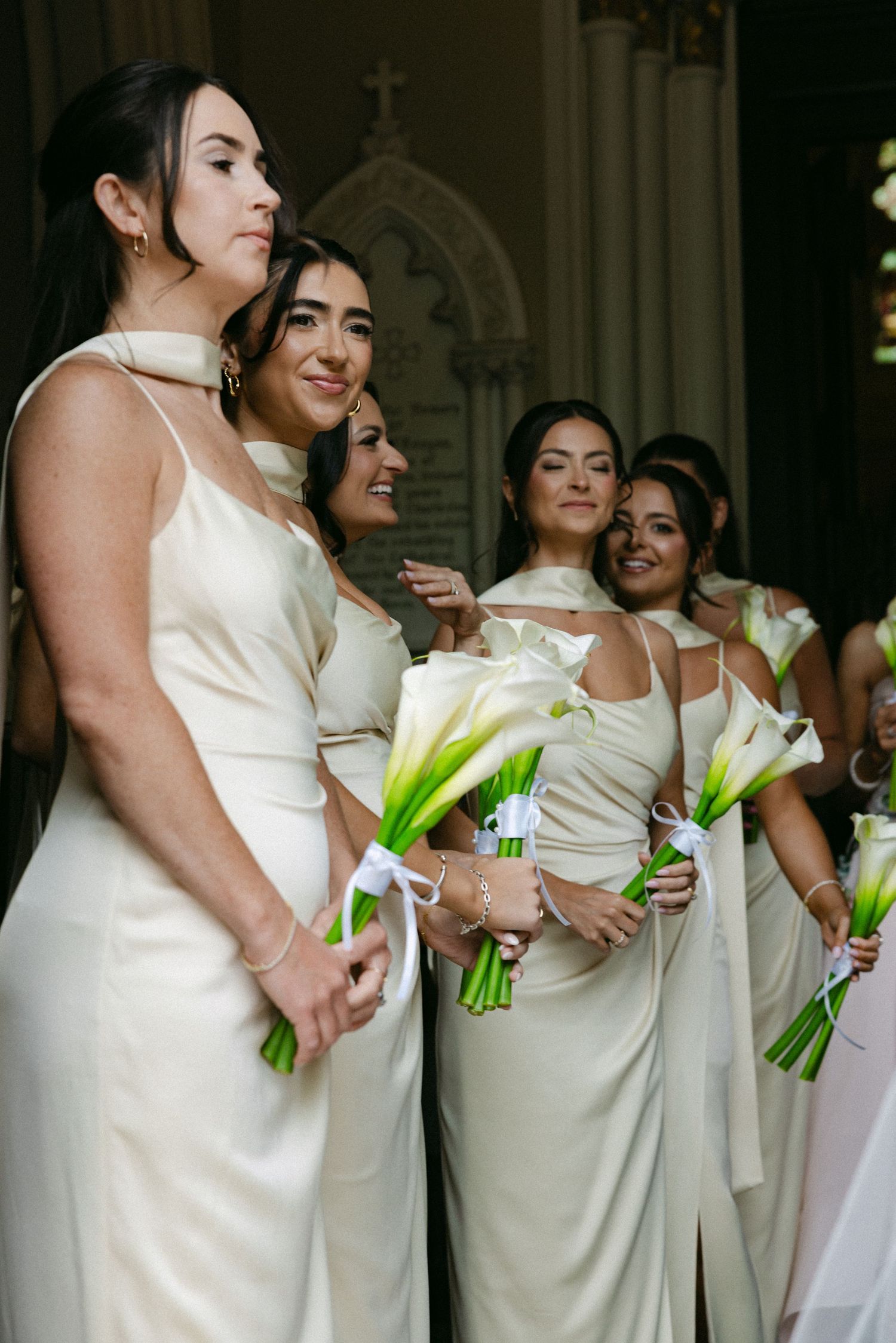 Bridesmaids in cream dresses hold green calla lily bouquets during church ceremony.