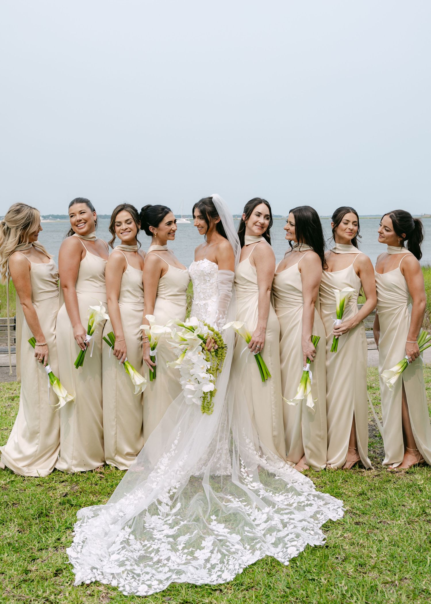 Bridal party with bridesmaids in champagne dresses lined up on grass with white floral bouquets.