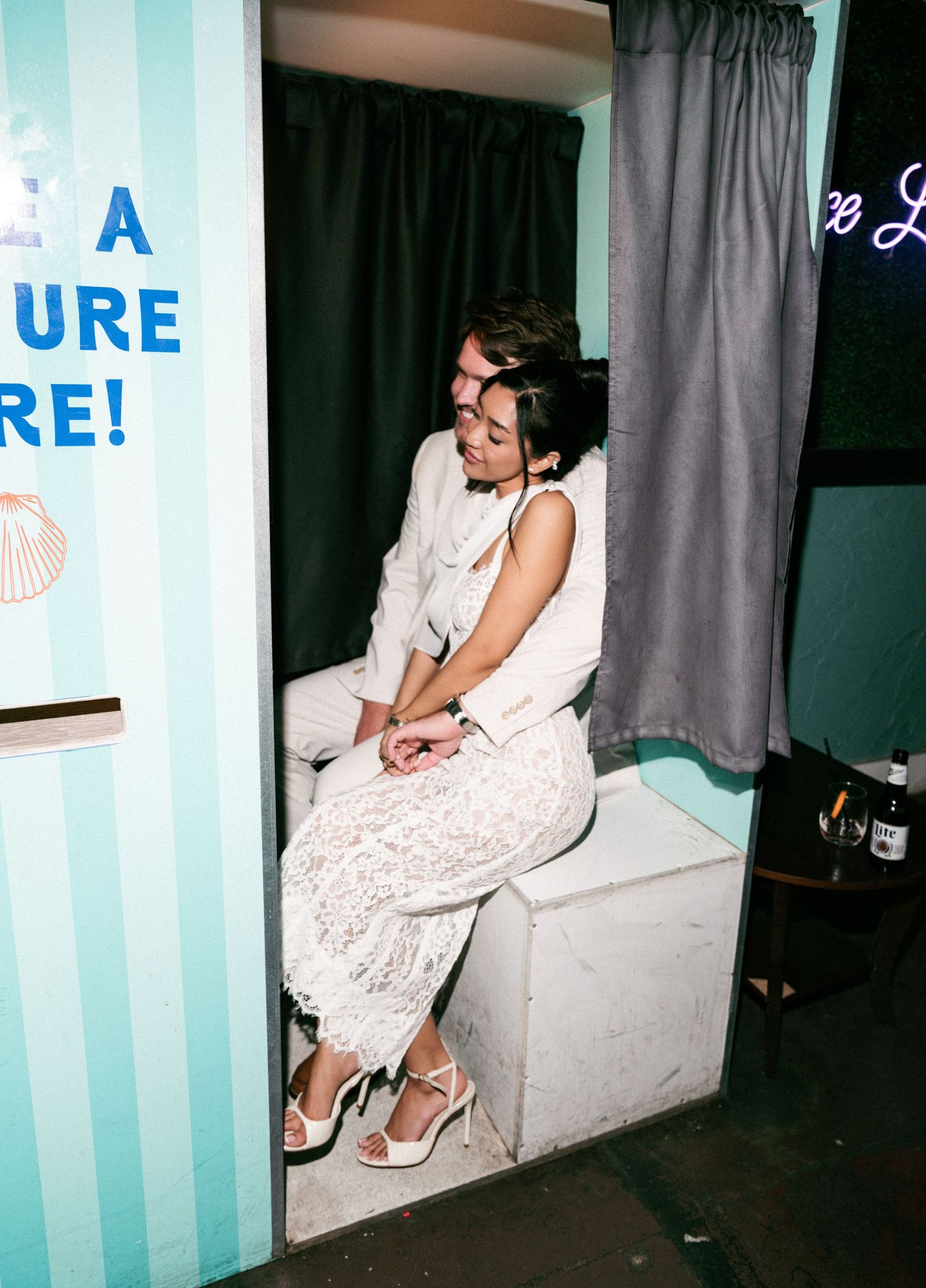 Couple sitting together in a vintage photo booth with gray curtain at a wedding reception event.
