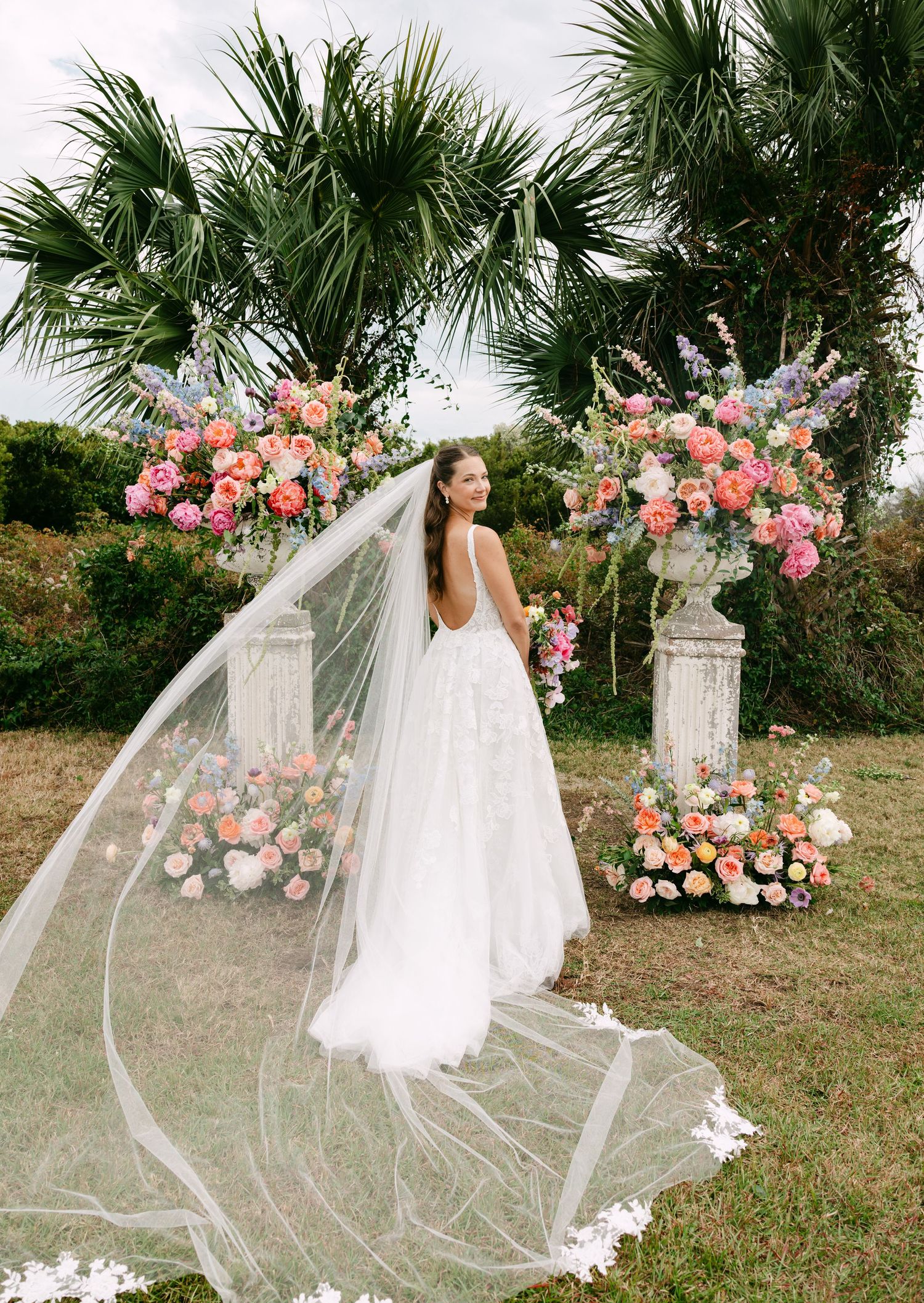 Bride in backless lace gown with cathedral veil posing before colorful floral pedestals outdoors.