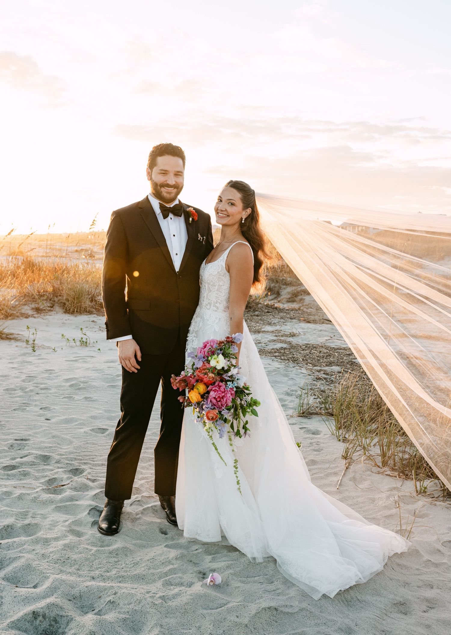 Bride and groom portrait on sandy beach at sunset, veil flowing in wind with colorful bridal bouquet.
