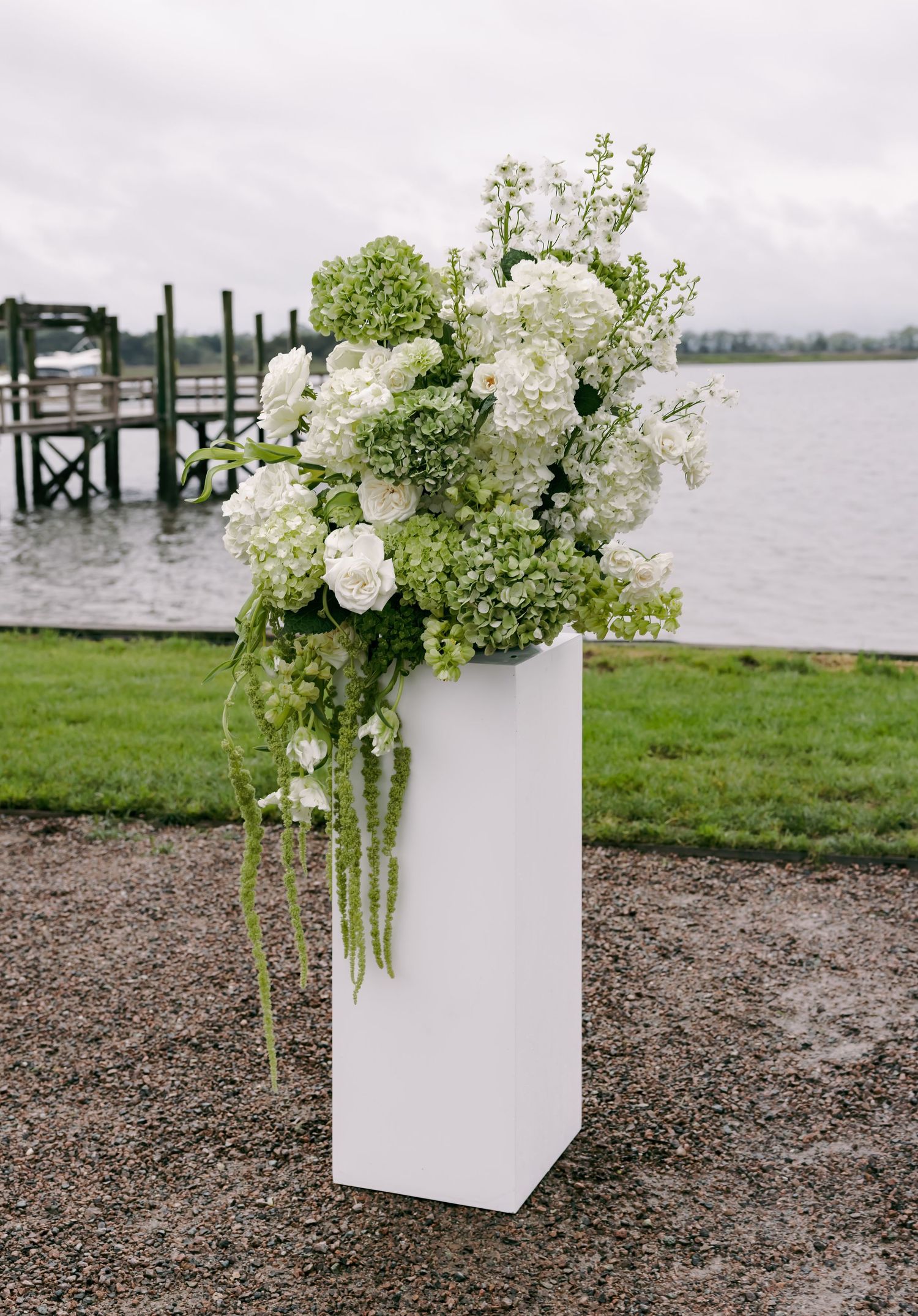 Lush white and green floral arrangement on white pedestal near waterfront dock at outdoor wedding ceremony.