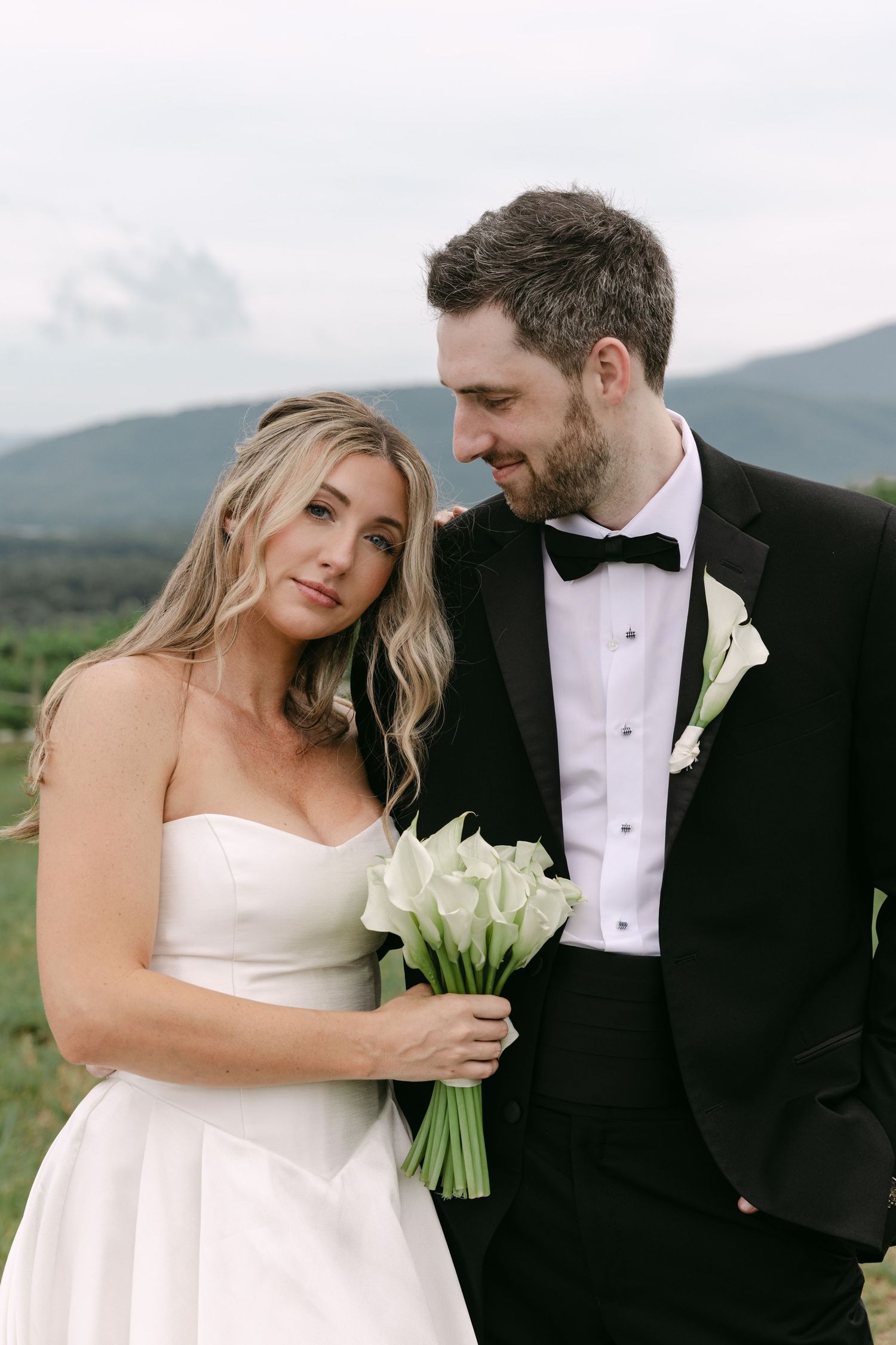 Bride holding white calla lily bouquet leans on groom in black tuxedo with mountain scenery behind them.