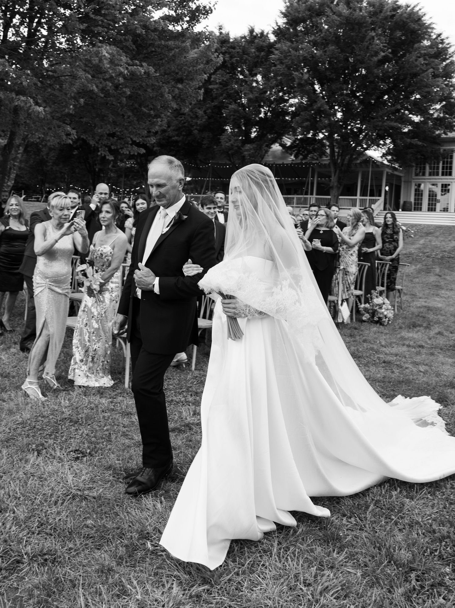 Bride in white gown and veil walks down aisle with escort at outdoor wedding ceremony.