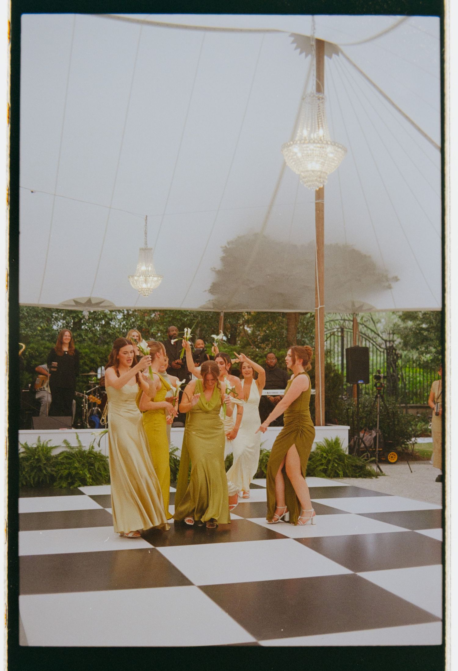 Bride and bridesmaids in green and gold dresses dancing on black and white checkered floor under chandelier tent.