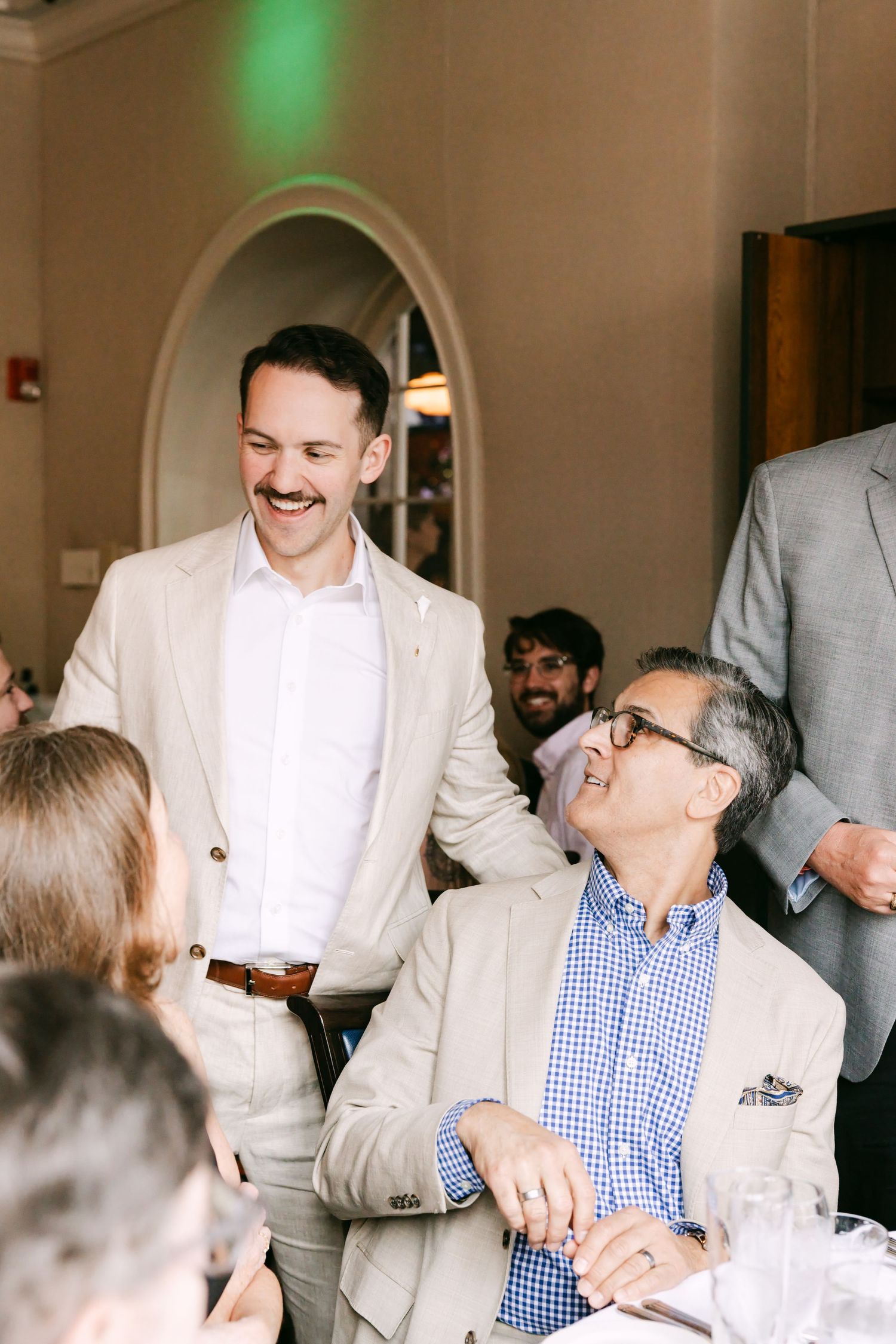Smiling groom in beige suit interacting with seated older gentleman in checkered shirt at wedding reception.