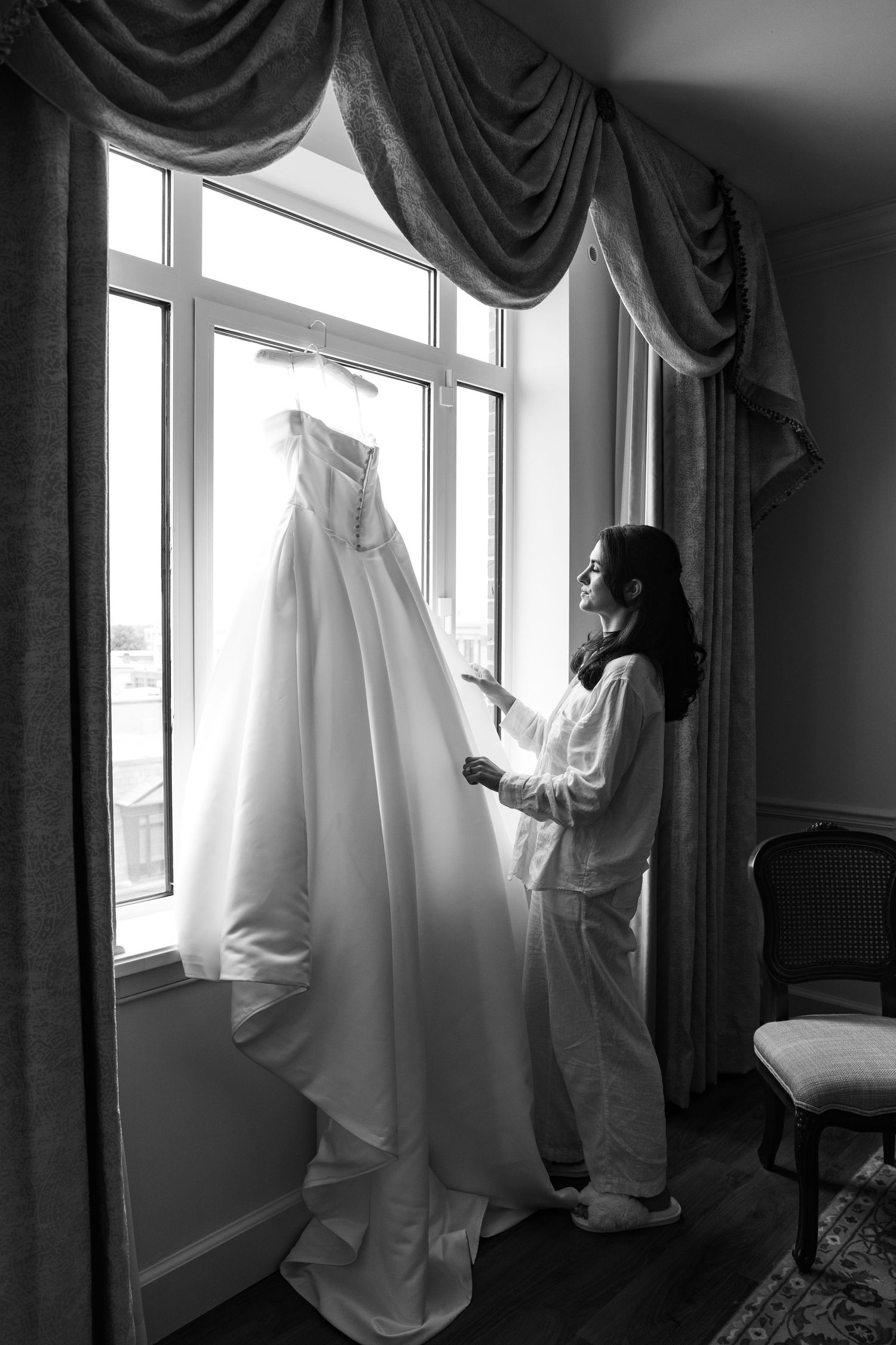 Bride in white robe admiring her wedding gown hanging by a bright window in a black and white photo.