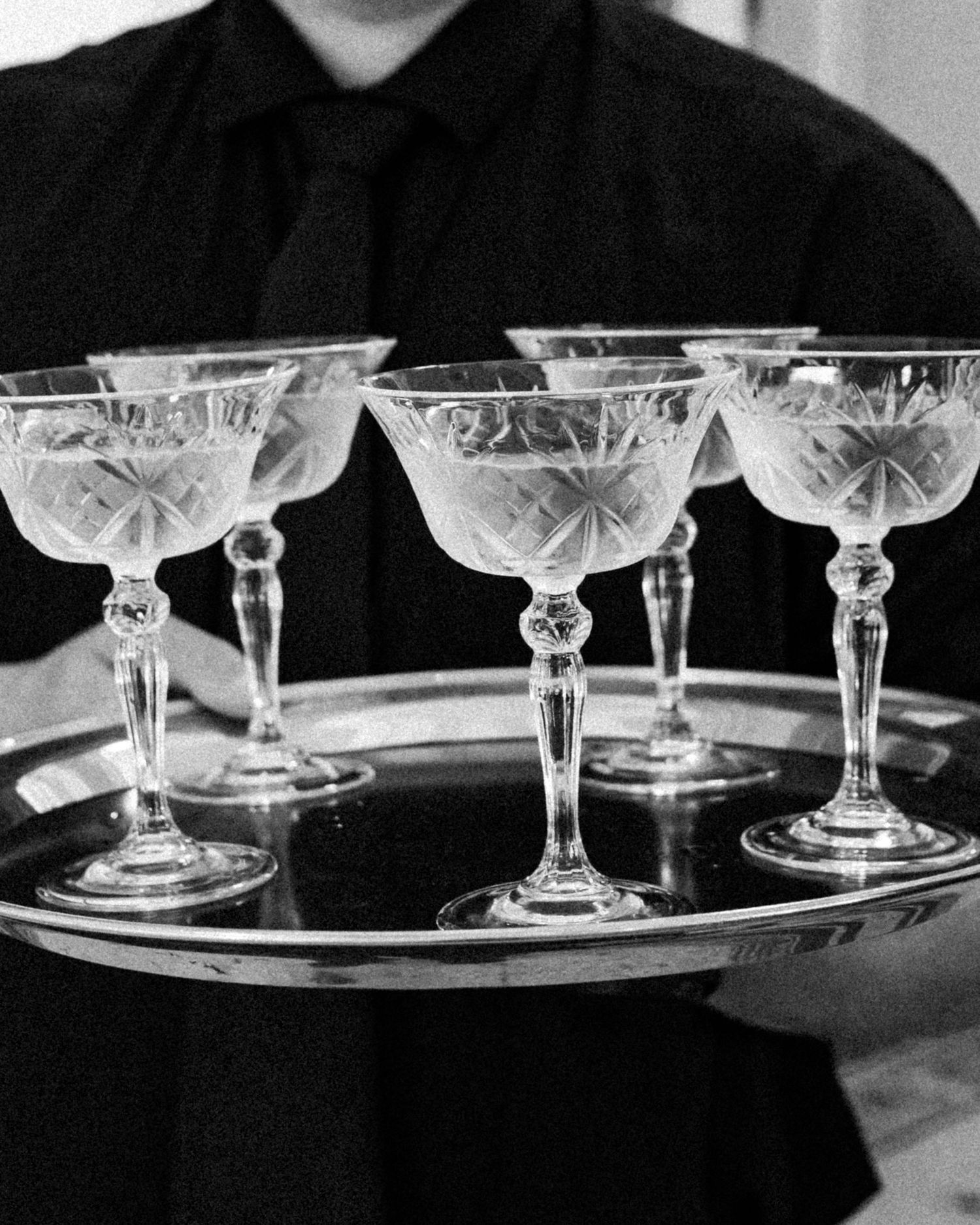 A waiter in black holds a silver tray with five elegant crystal coupe glasses in a black and white photo.