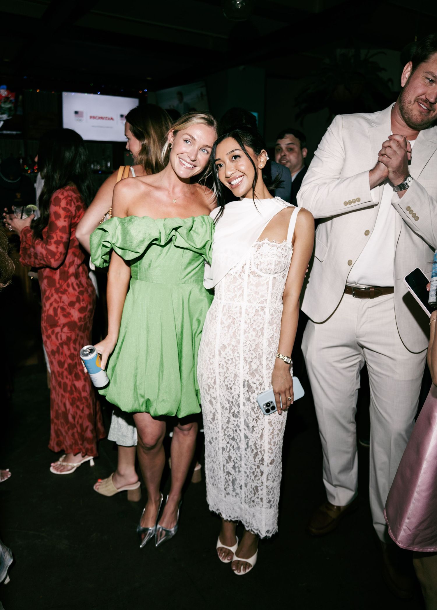 Two women smiling at a formal event, one in a green ruffled mini dress and one in a white lace midi dress.