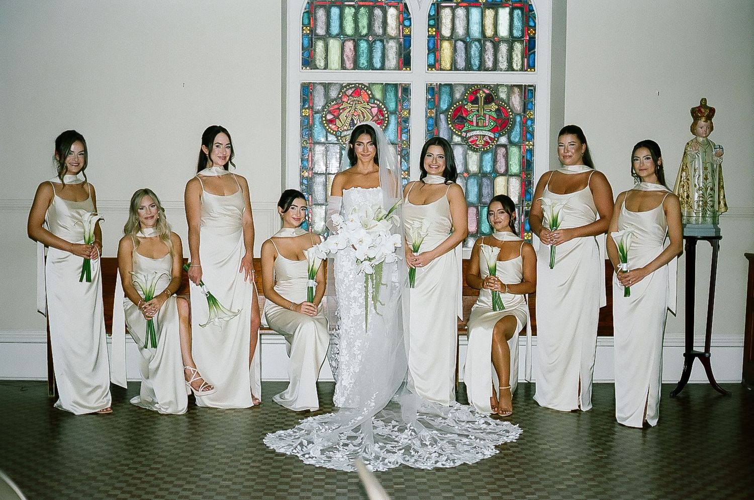 Wedding party in white dresses poses in front of stained glass windows.