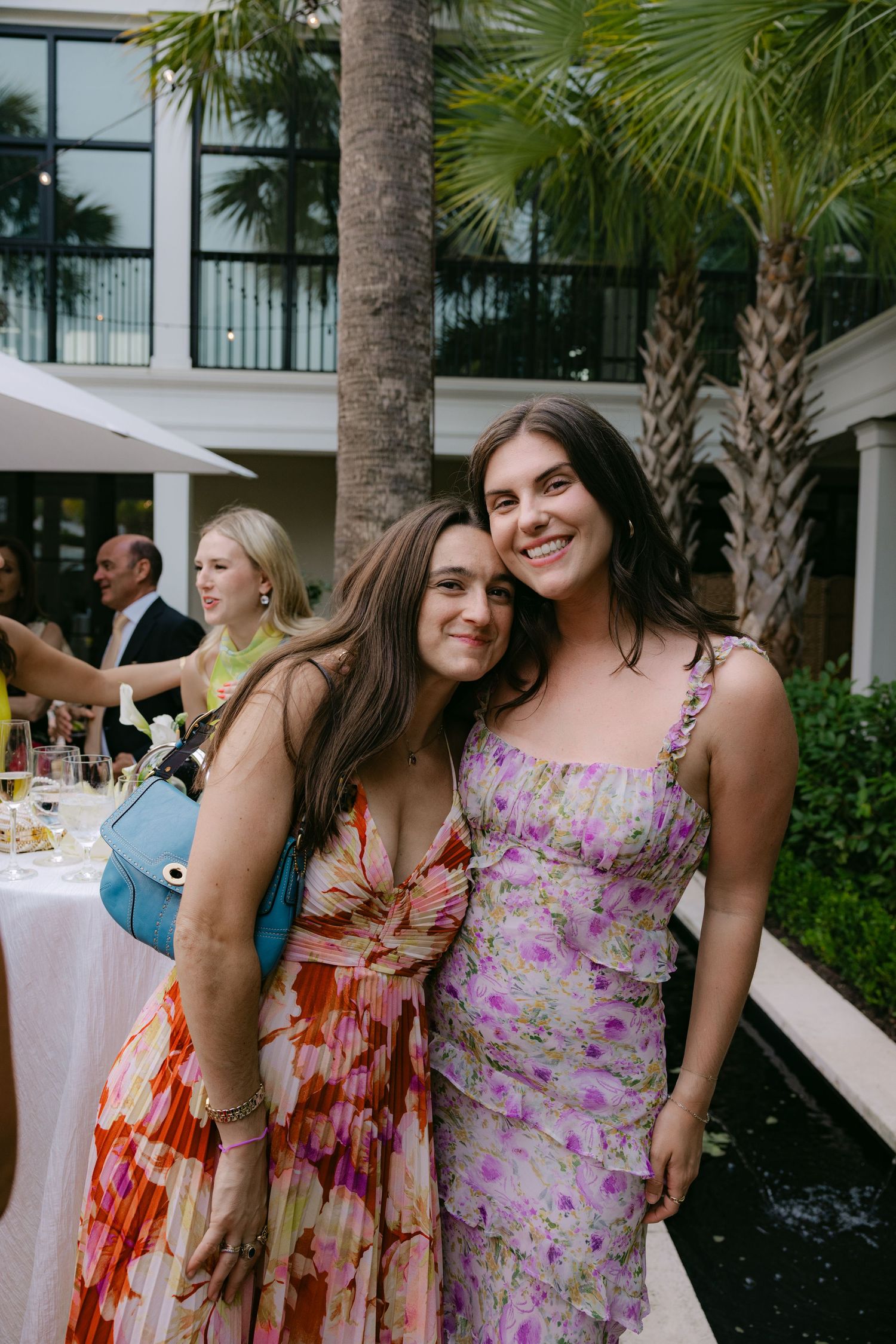 Bridesmaids in colorful floral dresses pose together at an outdoor wedding reception with palm trees in background.
