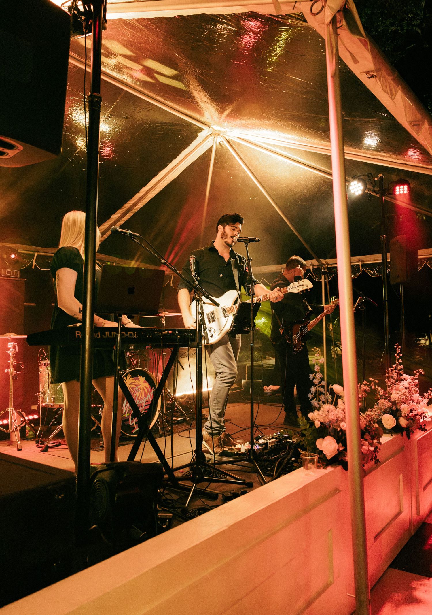 Live band performing under a tent with warm lights, guitarist smiling on stage with flowers and keyboard player nearby.