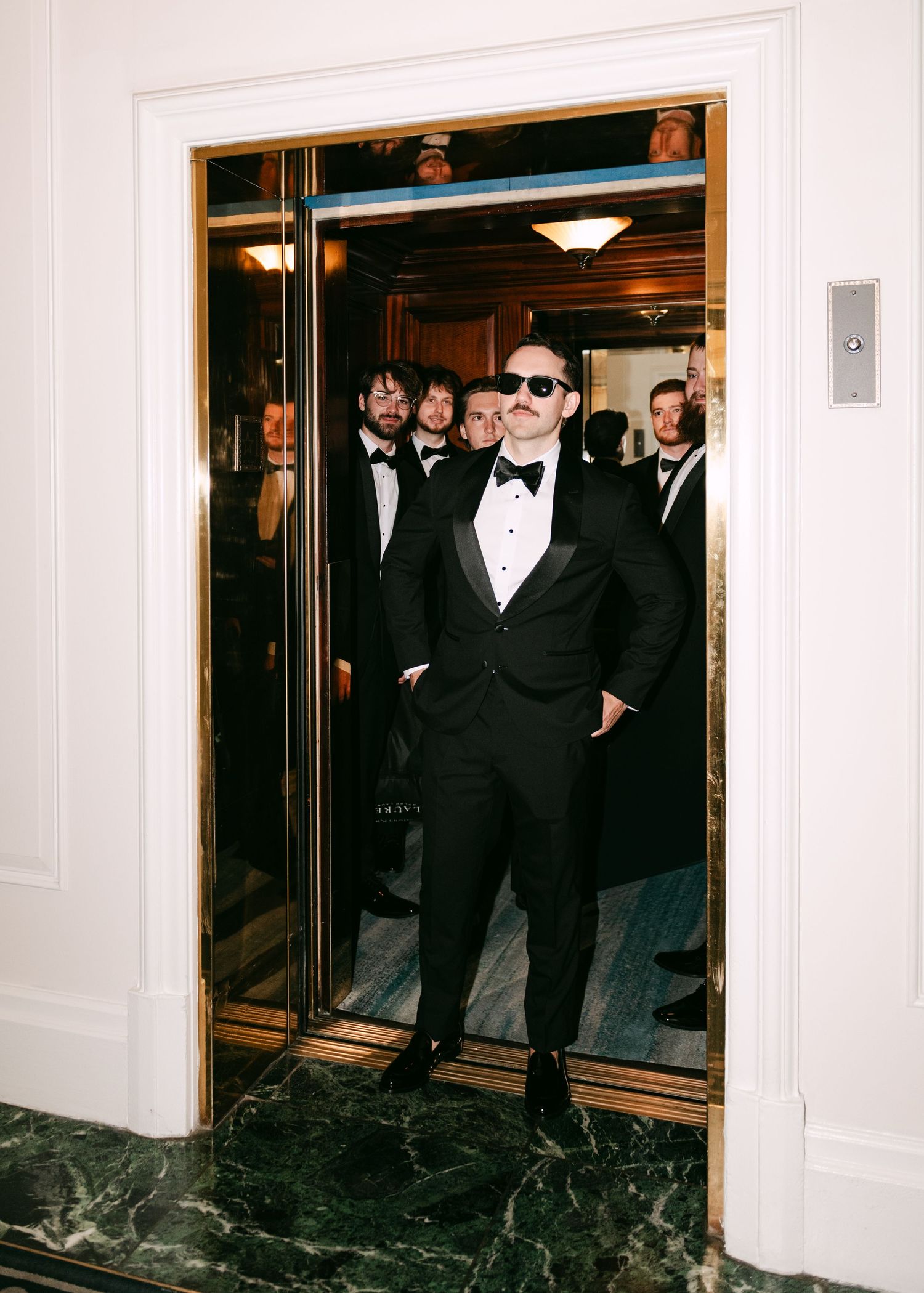 Groom in black tuxedo and sunglasses exits elevator with groomsmen at elegant wedding venue.